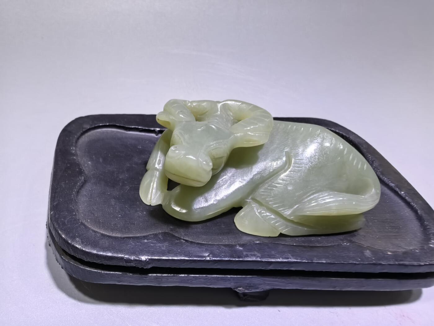 旧藏和田青白玉卧牛镇纸，传世雅物。
玉质青润细腻，包浆醇厚自然，雕工娴熟流畅，卧牛造型憨态可掬，栩栩如生。
牛角弯转，神态悠然，肌理线条精准传神，尽显古玉之韵与匠人之心。
既是文房镇纸，亦是案头雅赏，承载着“牛气冲天、扭转乾坤”的美好寓意，为藏家不可多得的臻品。5.5cm＊3.6cm＊2.0cm
克重：53.2g
