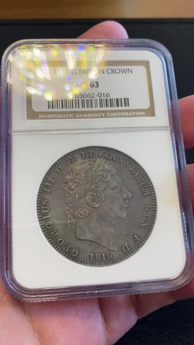 万国钱币拍卖第005期 1819LIX英国乔治三世马剑银币，英国马剑系列的开端，机制克朗的鼻祖，也代表着蒸汽时代的到来。马剑图案由意大利雕刻家Pistrucci设计，在钱币两面皆留下了显而易见的签名，这在当时是难以被接受的（一般只会留下首字母缩写），此举也为后来首席雕刻师W.Wyon的上位埋下了伏笔。此枚钱币高点细节皆属完美，略带镜面感的底板在光线下呈现出五彩斑斓的色彩，整体状态有着不输64分的水准。
