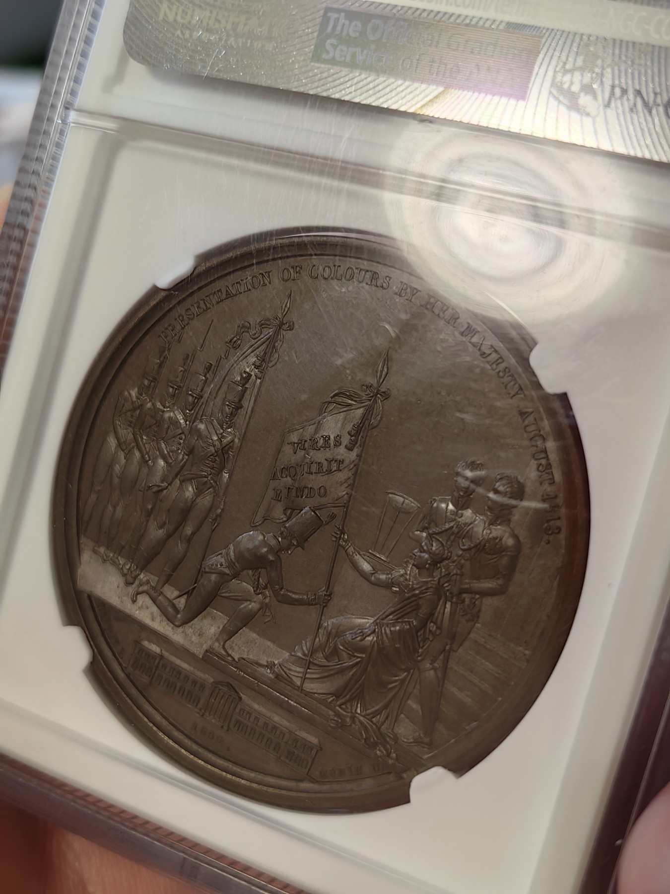 橡树集藏-006场 海纳百川 1813年英国皇室章，乔治三世时期原铸。ngc Ms65，加厚盒。铜色诱人，镜面感十足。背面是女王向皇家近卫军团授予军旗，细节丰富，雕刻精细，超高浮雕。ngc数据库唯一评级，冠军分。军旗上刻字：VIRES ACQUIRT EUNDO。拉丁文，意为“随行聚力”，是古罗马诗人的叙事诗《埃涅阿斯纪》中的一句，意思是“随着传播而越来越有力”。象征着军队听从指挥，雷厉风行。