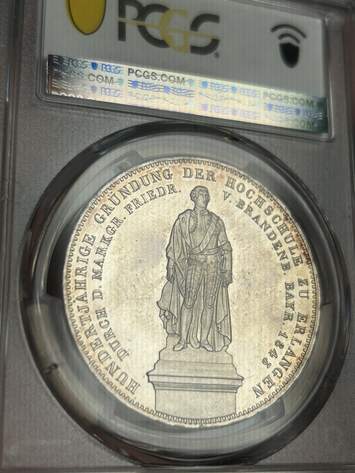 万国钱币拍卖第021期（龙年首拍） PCGS MS64 1843巴伐利亚埃尔朗根大学建校百年历史2泰勒 背面为大学创始人勃兰登堡-拜罗伊特边疆伯爵腓特烈的雕像，相当于中国古代的藩王。此枚品相状态已属顶级，镜面感极强的底板在双面淡金彩的加持下熠熠生辉，腓特烈伯爵的雕像细节一丝不苟凝于方寸之间，数百年之后辗转腾挪来到诸位手中，可能这也就是路德维希一世铸造历史的用意所在。