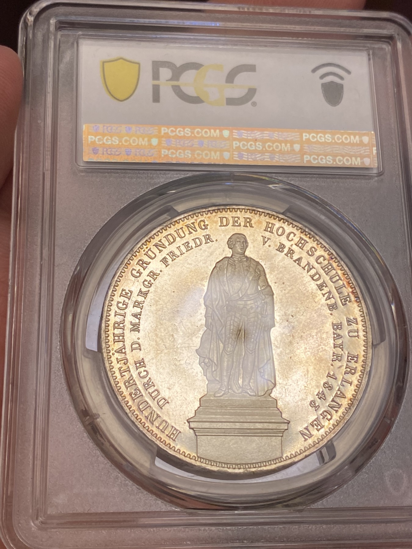 万国钱币拍卖第021期（龙年首拍） PCGS MS64 1843巴伐利亚埃尔朗根大学建校百年历史2泰勒 背面为大学创始人勃兰登堡-拜罗伊特边疆伯爵腓特烈的雕像，相当于中国古代的藩王。此枚品相状态已属顶级，镜面感极强的底板在双面淡金彩的加持下熠熠生辉，腓特烈伯爵的雕像细节一丝不苟凝于方寸之间，数百年之后辗转腾挪来到诸位手中，可能这也就是路德维希一世铸造历史的用意所在。
