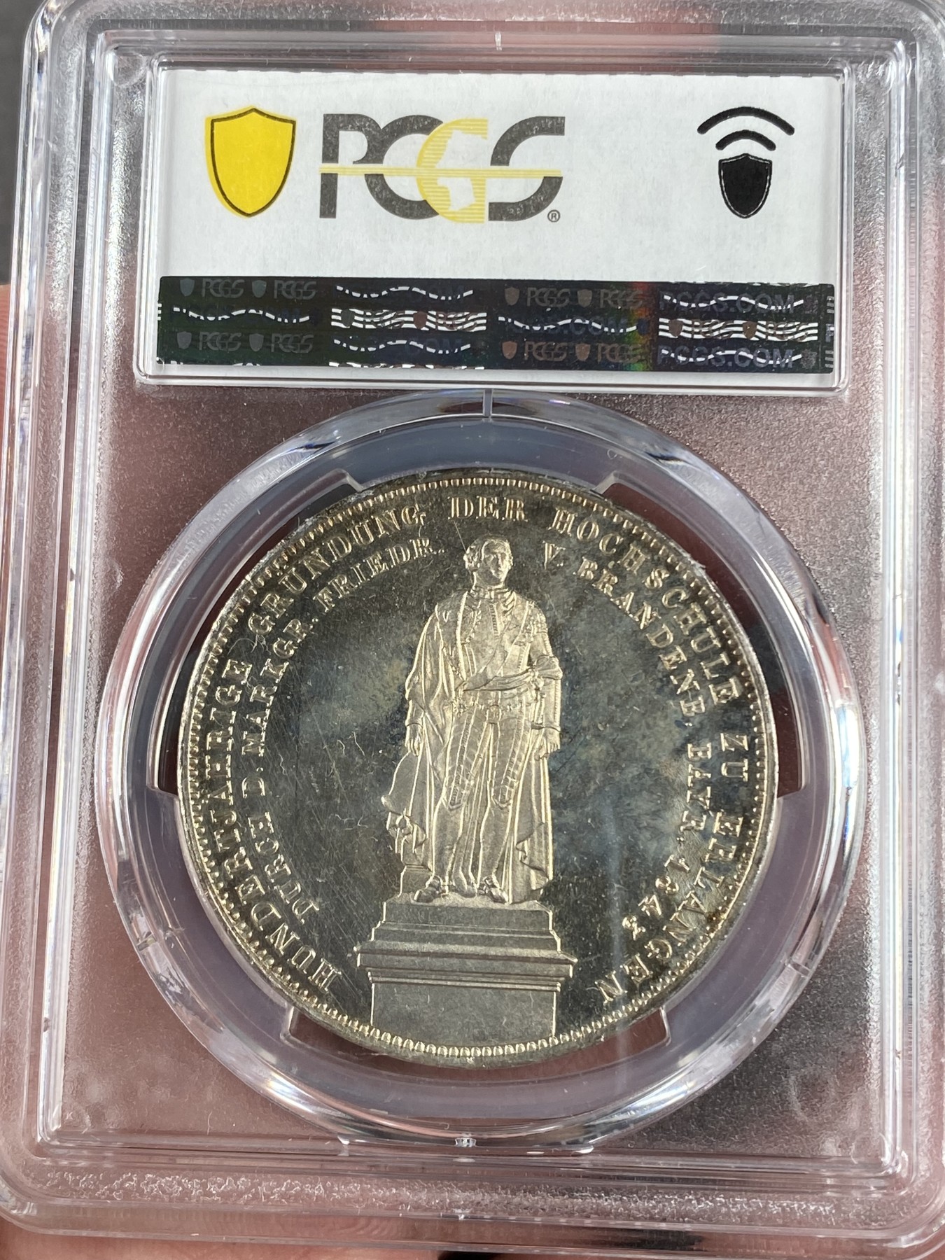 万国钱币拍卖第021期（龙年首拍） PCGS MS64 1843巴伐利亚埃尔朗根大学建校百年历史2泰勒 背面为大学创始人勃兰登堡-拜罗伊特边疆伯爵腓特烈的雕像，相当于中国古代的藩王。此枚品相状态已属顶级，镜面感极强的底板在双面淡金彩的加持下熠熠生辉，腓特烈伯爵的雕像细节一丝不苟凝于方寸之间，数百年之后辗转腾挪来到诸位手中，可能这也就是路德维希一世铸造历史的用意所在。