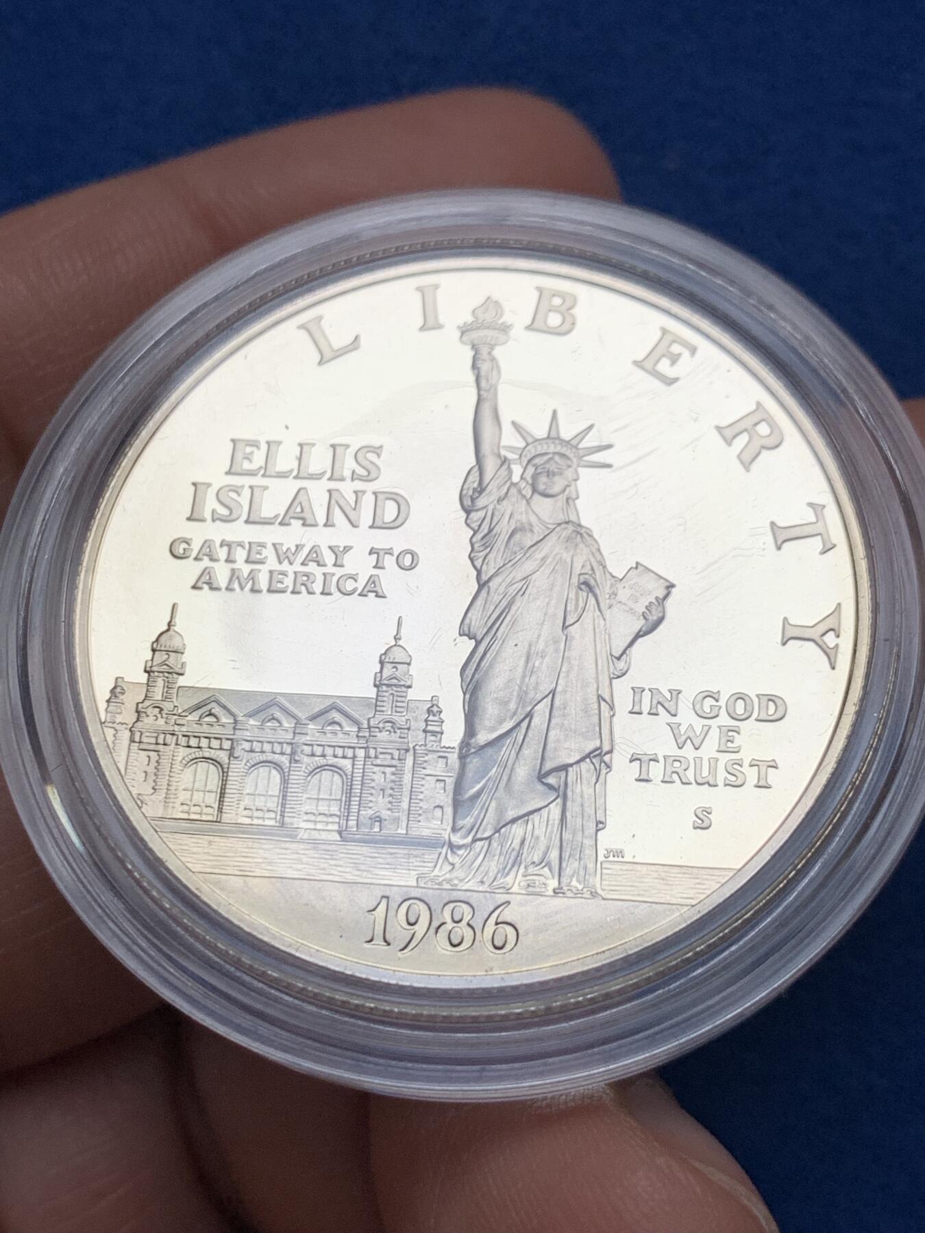 《竞宝斋》第353场 周日，周一，周二 3场连拍 （全场包邮） 美国1986年纪念自由女神像建成100周年精制银币套装。银币正面为庄严肃穆自由女神像，银币反面为自由女神像的火炬以及诗歌《新巨人》中的文字。 半美元铜镍币正面为纽约的天际线，自由女神像举起手臂，迎接一艘驶向纽约的班轮，太阳从东方升起，寓意着新世界新生活的开始。背面图案源自埃利斯岛上等待前往纽约的移民照片，反映了自由女神像作为移民希望象征的意义。