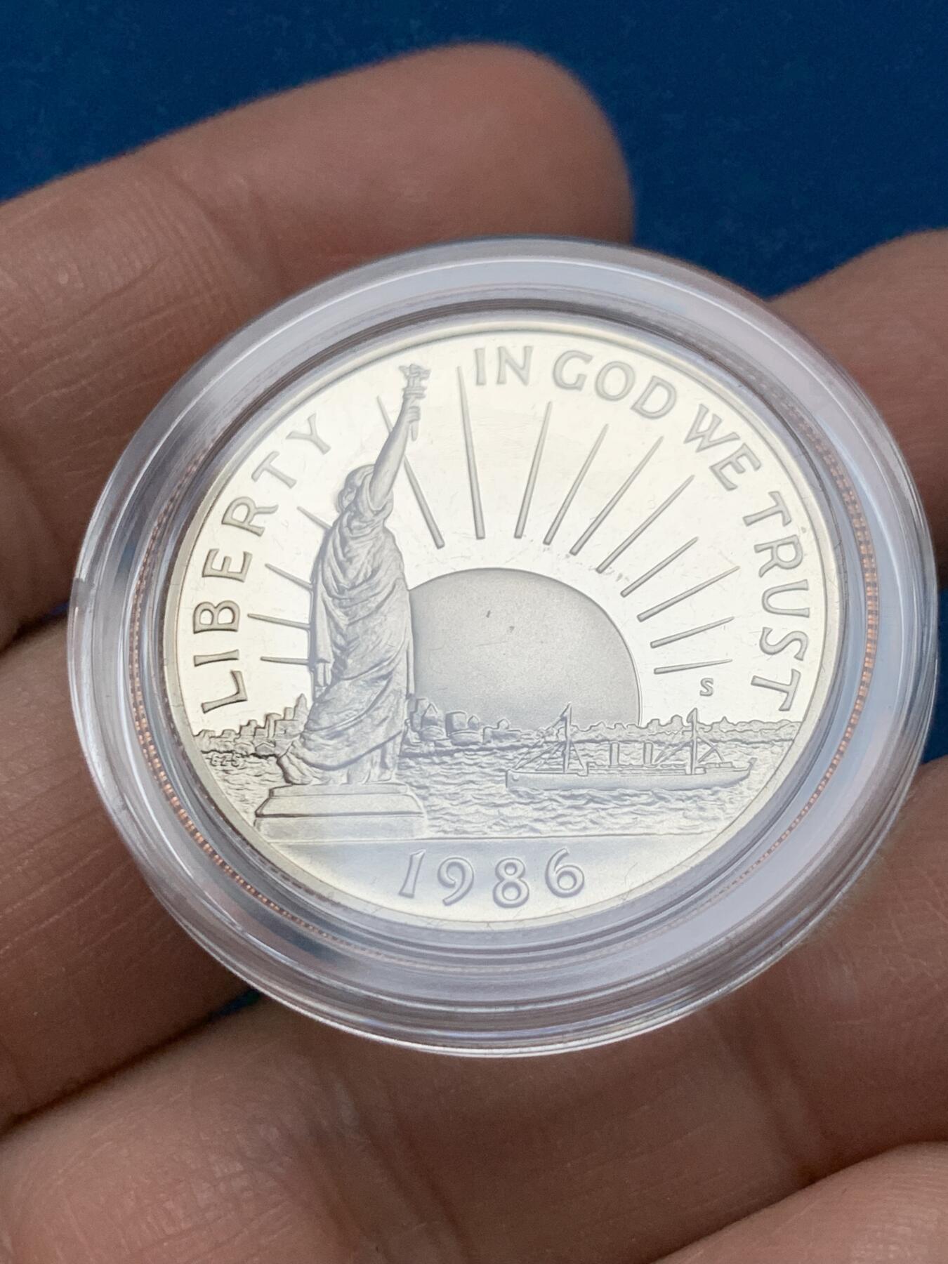 《竞宝斋》第353场 周日，周一，周二 3场连拍 （全场包邮） 美国1986年纪念自由女神像建成100周年精制银币套装。银币正面为庄严肃穆自由女神像，银币反面为自由女神像的火炬以及诗歌《新巨人》中的文字。 半美元铜镍币正面为纽约的天际线，自由女神像举起手臂，迎接一艘驶向纽约的班轮，太阳从东方升起，寓意着新世界新生活的开始。背面图案源自埃利斯岛上等待前往纽约的移民照片，反映了自由女神像作为移民希望象征的意义。