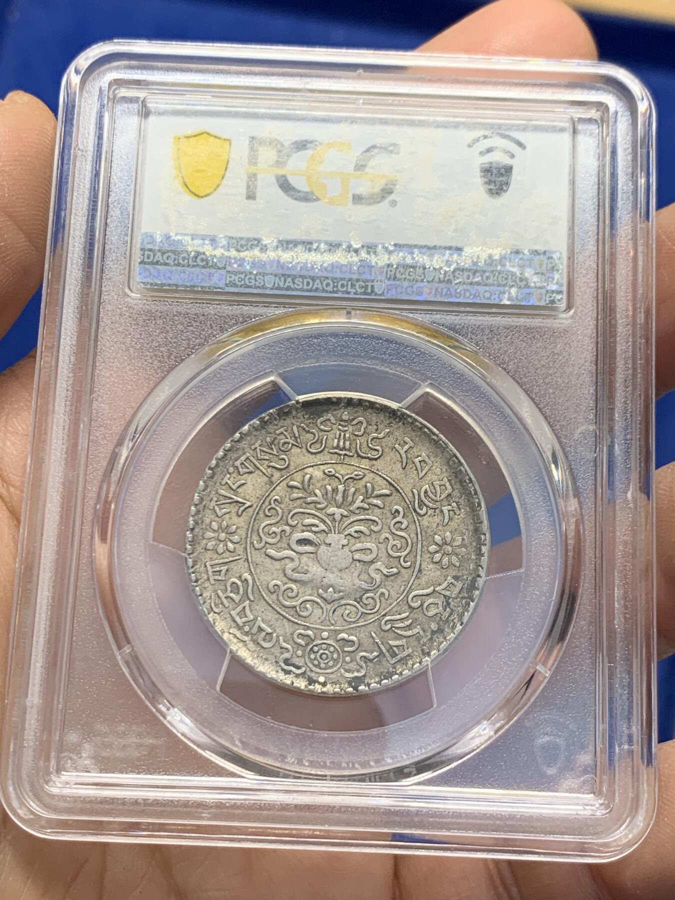 《竞宝斋》第353场 周日，周一，周二 3场连拍 （全场包邮） PCGS AU55 中国1937年西藏桑松果木三两银币，正面中央圆环内为日月、冈仁波齐雪山、祥云、狮子、绣球图案，外圈由宝伞（左上）、宝鱼（右上）、白海螺（右下）、吉祥结（左下）作为间隔环刻藏文“甘丹颇章，战胜四方”字样。背面中央圆环内为花枝和宝瓶图案，周围环绕缠枝纹和飘带；外圈以胜利幢（上）、金法轮（下）和莲花（左右）作为间隔，环刻藏文纪年、面值。具有独特的历史背景、文化内涵和艺术特征。此枚原味