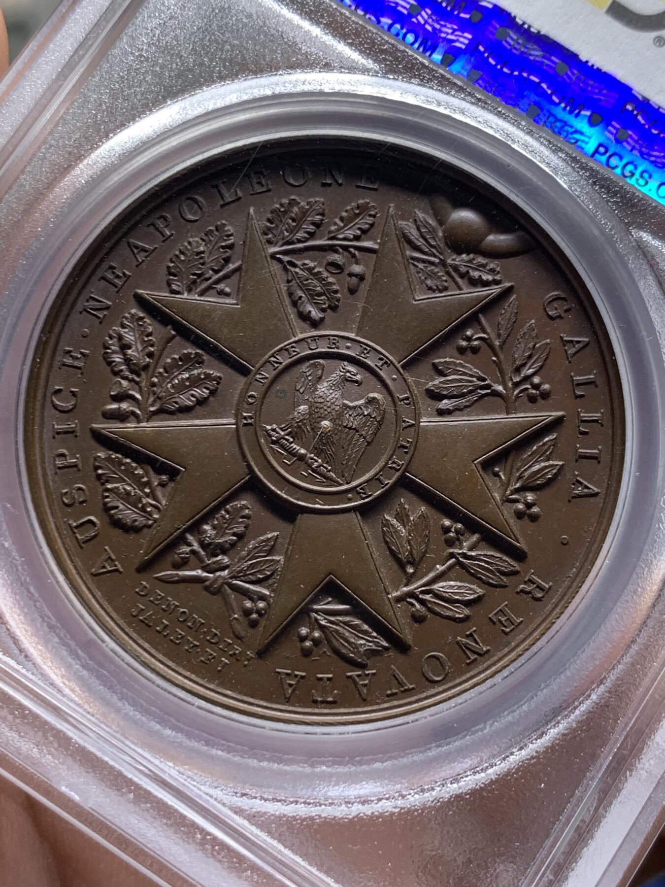 《竞宝斋》第387场 周日，周一 2场连拍 （全场包邮） PCGS SP65 冠军分 法国1804年拿破仑荣誉军团原铸铜章 巴黎造币厂巧克力包浆 荣誉军团勋章交织在月桂树和橡树花环间 ，创造性地将这样一枚传奇的勋章呈现于铜章之上，创意十足远超时代，上部佩环，中间是拿破仑鹰徽和该勋章的铭文 Honneur et Patrie（“荣誉和祖国”），是法国最高的功绩勋章，由拿破仑本人于1802年创立，高浮雕品相顶级 ，属于拿章中的稀少名誉品种。加厚盒。