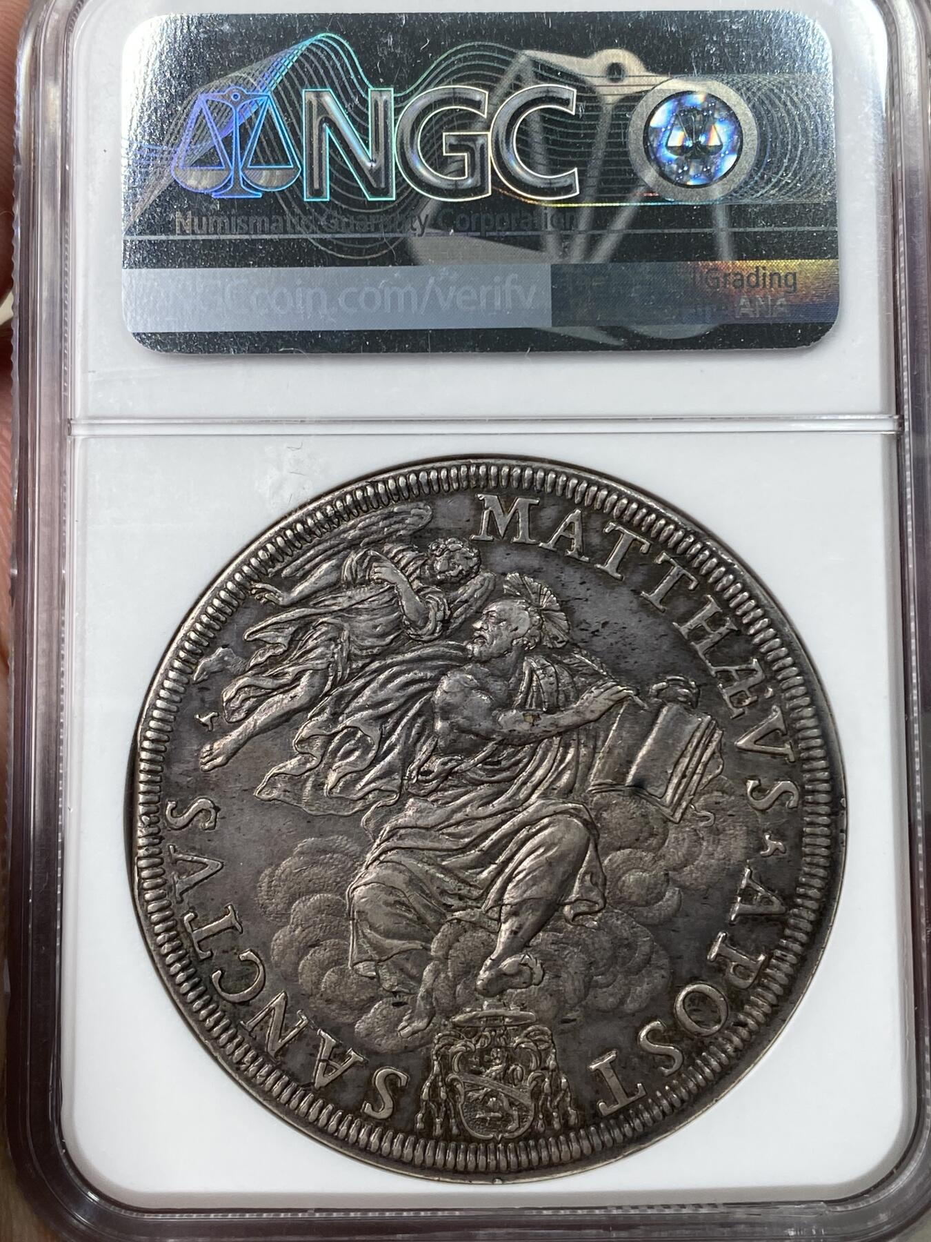 万国钱币拍卖第049期（万国夏季大拍） NGC AU58 1676年意大利教皇国因诺森十一世1皮阿斯特大银，正面为身着繁复华丽教宗服饰的因诺森十一世，背面为拿着书本的圣马太和天使，早期教皇国任何大银都极其昂贵，58已是佼佼者。此枚灰黑色均匀包浆，打制深峻，教皇国玩家不容错过。