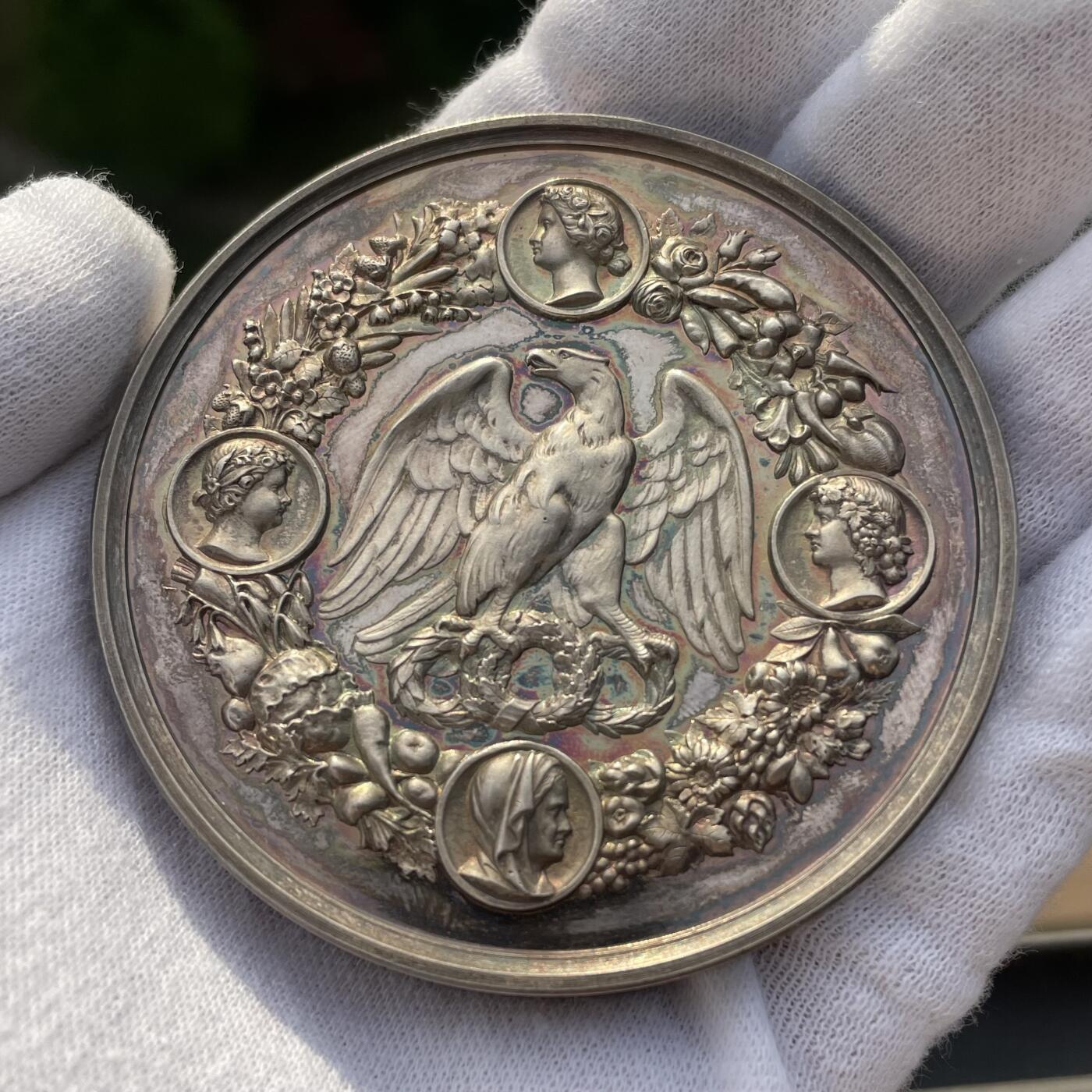 「木头集藏」章牌精品专场！ Mertens & Schilling～1861年德国园艺银质奖章，直径：65mm，120.8 g，普鲁士官方颁发的园艺成就奖。左侧为坐着的女性形象，手持丰饶角，左侧则有一把耙子、一个洒水壶，以及一棵环绕着花果花环的小树/老鹰，上方为四个象征四季的女性头像徽章，非常漂亮的包浆和品相。