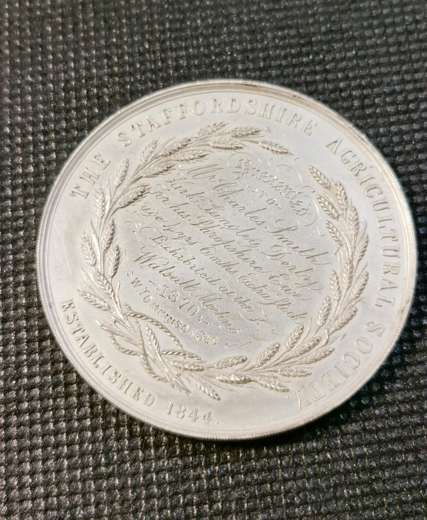  甄臻铺-世界币章拍卖（第24期），送拍享返佣金等优惠多多 1870年斯塔福德郡农业协会颁发的银质奖章，正面刻有农场动物群雕，包括马、牛、猪、鸡等，背景是农舍和教堂塔楼，雕刻精美生动。背面刻有协会名称、成立年份1844年，以及获奖者Charles Smith的铭文，环绕麦穗花环装饰。奖章直径约47.2毫米，重31.3克，保存完好，表面有轻微使用痕迹，适合收藏。这枚奖章是当时农业展示的重要纪念品，具有历史研究价值。
