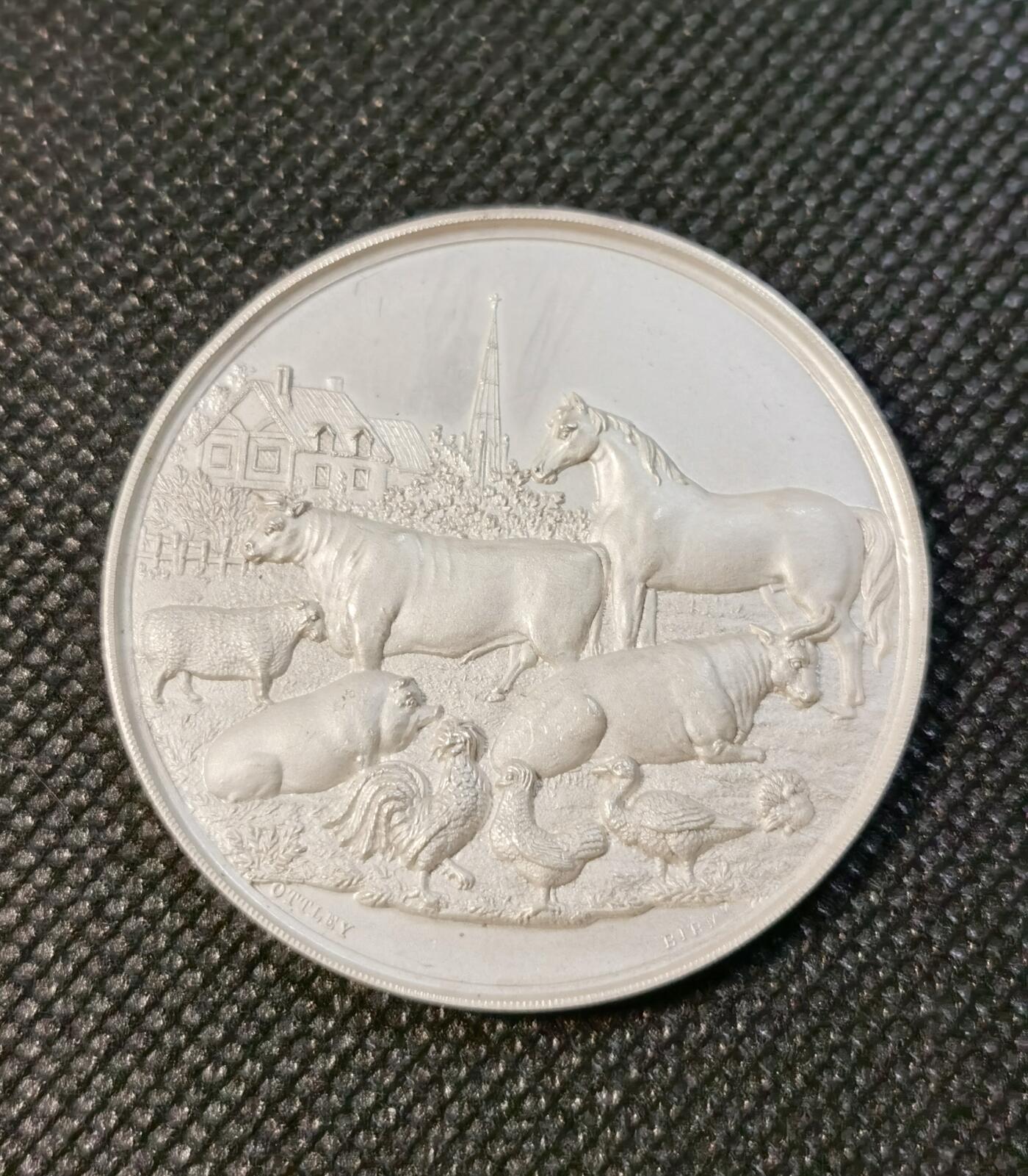  甄臻铺-世界币章拍卖（第24期），送拍享返佣金等优惠多多 1870年斯塔福德郡农业协会颁发的银质奖章，正面刻有农场动物群雕，包括马、牛、猪、鸡等，背景是农舍和教堂塔楼，雕刻精美生动。背面刻有协会名称、成立年份1844年，以及获奖者Charles Smith的铭文，环绕麦穗花环装饰。奖章直径约47.2毫米，重31.3克，保存完好，表面有轻微使用痕迹，适合收藏。这枚奖章是当时农业展示的重要纪念品，具有历史研究价值。