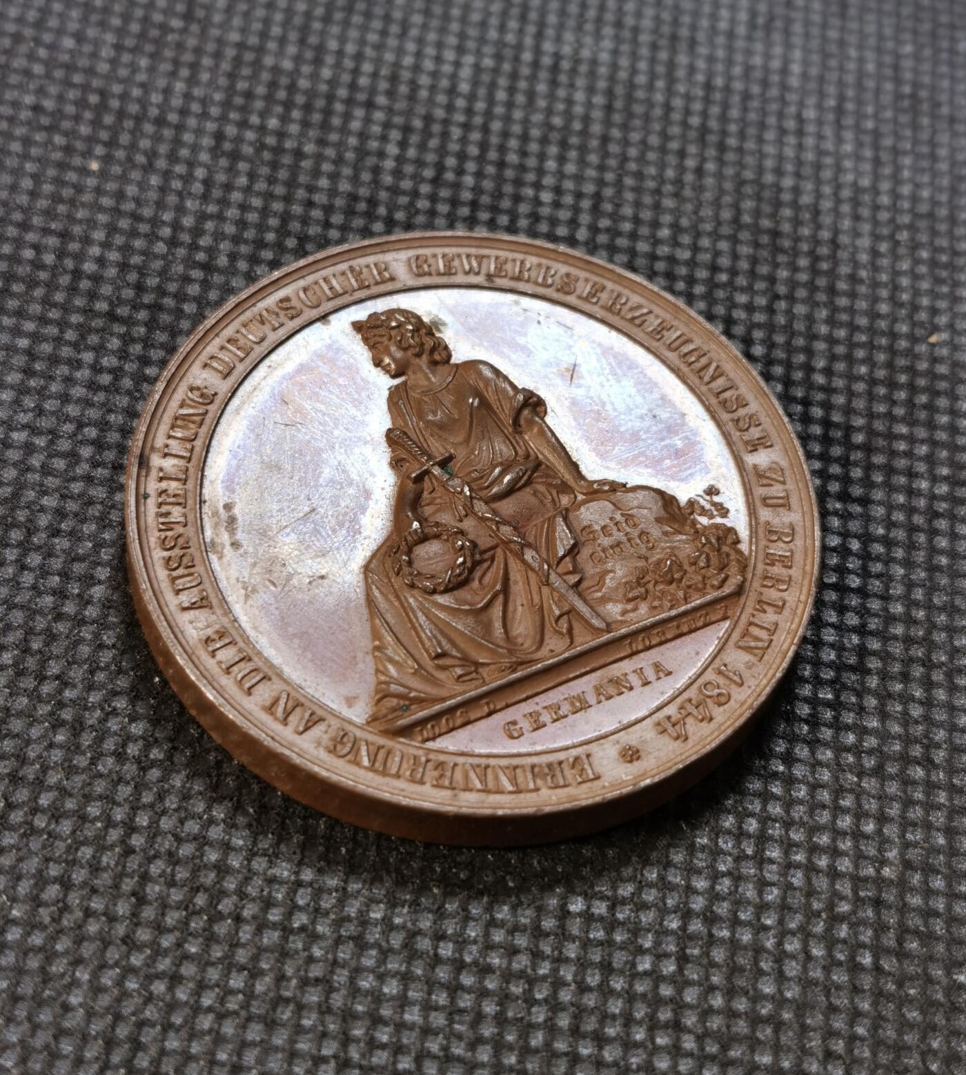  甄臻铺-世界币章拍卖（第24期），送拍享返佣金等优惠多多 1844年德国柏林工业博览会纪念铜章，正面是德意志女神Germania的半身像，背面展示工业成就场景，包括蒸汽机车和其他工业设备。由德国雕刻家创作，直径45毫米，重53.89克。铜质精良，雕工细腻，历史意义重大。正面德文铭文完整，背面环绕工业主题装饰纹样，体现了当时德国工业发展的繁荣景象