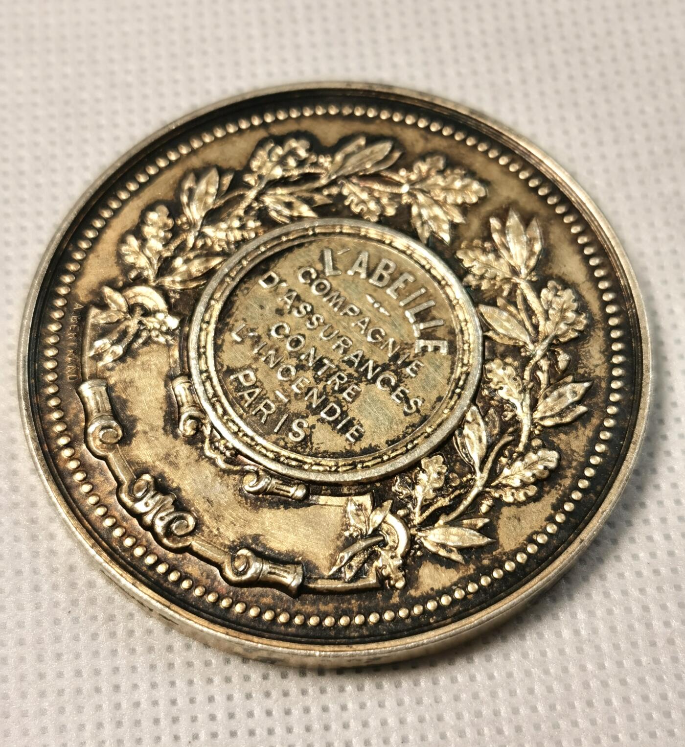  甄臻铺-世界币章拍卖（第25期），送拍享返佣金等优惠多多 法国L'ABEILLE保险公司颁发的获奖镀金银章，直径50.7毫米，重65.3克。正面是雅典娜女神浮雕，手持橄榄枝，象征和平与保护。背面环绕橄榄枝花环，刻有公司名称