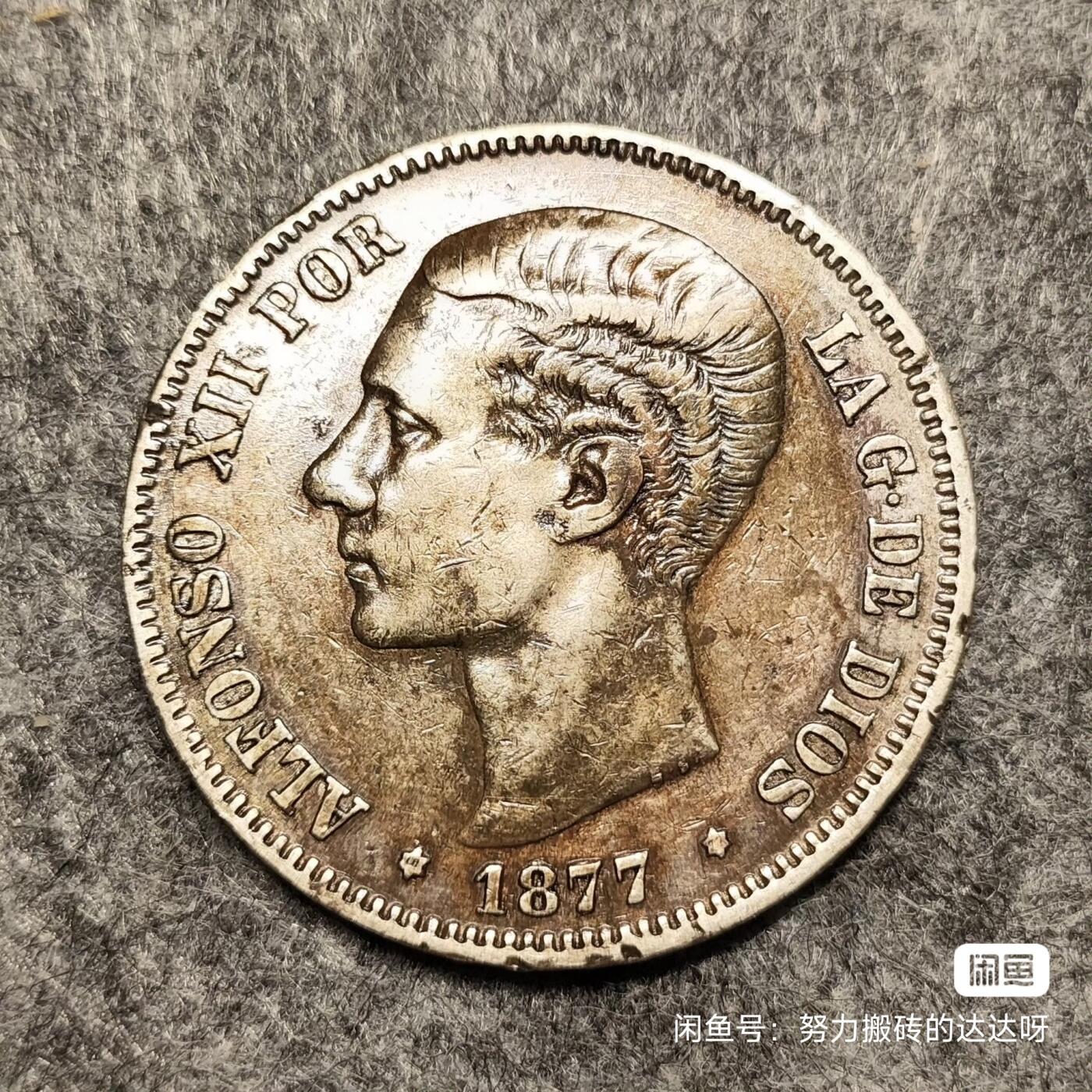 西班牙1877年阿方索十二世双柱5比塞塔银币 青年版少见。银币重25g，含银量90%。正面是西班牙国王阿方索十二世青年肖像，外环为拉丁文铭（阿方索十二世蒙神之恩）。背面图案是西班牙盾徽，盾徽中间椭圆内有三朵鸢尾花代表波旁王朝家族，椭圆旁有4栏分别代表四个王国。两侧各列一根海格力斯柱柱上卷轴绕裹，卷轴上有“海外有大陆”。头发细节全酱彩包浆，稀少版别流通好品。售出不退不换菜鸟包邮