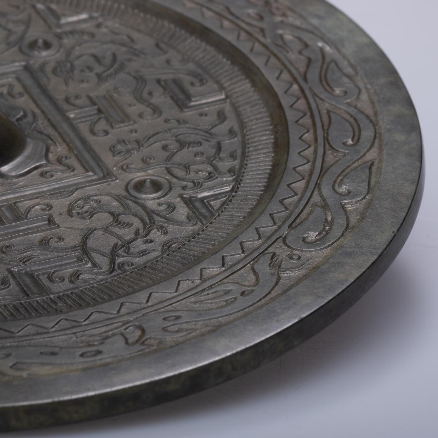 日本大阪雅和auction 2025金秋競賣會第一期 漢 走獸紋規矩鏡 尺寸:D:14cm 備註:盒付   此面漢代走獸紋規矩鏡，鏡體渾圓厚重，通體黑漆古包漿，色澤沉潤柔和，鏡背以精細線條鑄出走獸紋飾，或奔或躍，神態生動，間以幾何規矩紋相間佈局，構圖嚴謹而富節奏感。鏡紐高起，形制端正，邊廓勻整，展現漢代青銅鏡工藝的成熟與規範之美。此類鏡多寓意驅邪護身、吉祥長安，包漿溫潤，光澤深邃，鏡面保存完好，實屬難得。其兼具實用與審美價值，彰顯漢代金屬藝術的莊重典