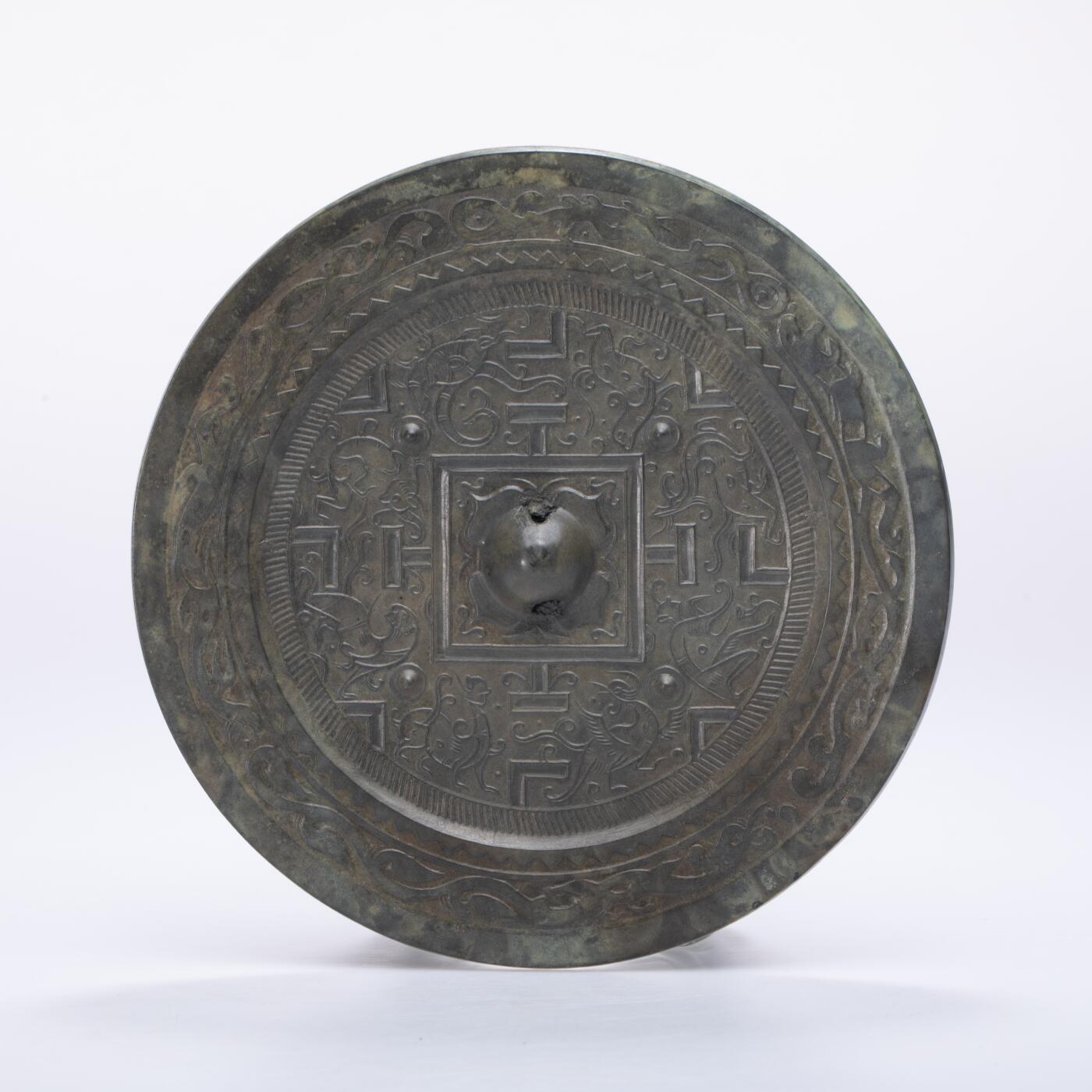 日本大阪雅和auction 2025金秋競賣會第一期 漢 走獸紋規矩鏡 尺寸:D:14cm 備註:盒付   此面漢代走獸紋規矩鏡，鏡體渾圓厚重，通體黑漆古包漿，色澤沉潤柔和，鏡背以精細線條鑄出走獸紋飾，或奔或躍，神態生動，間以幾何規矩紋相間佈局，構圖嚴謹而富節奏感。鏡紐高起，形制端正，邊廓勻整，展現漢代青銅鏡工藝的成熟與規範之美。此類鏡多寓意驅邪護身、吉祥長安，包漿溫潤，光澤深邃，鏡面保存完好，實屬難得。其兼具實用與審美價值，彰顯漢代金屬藝術的莊重典