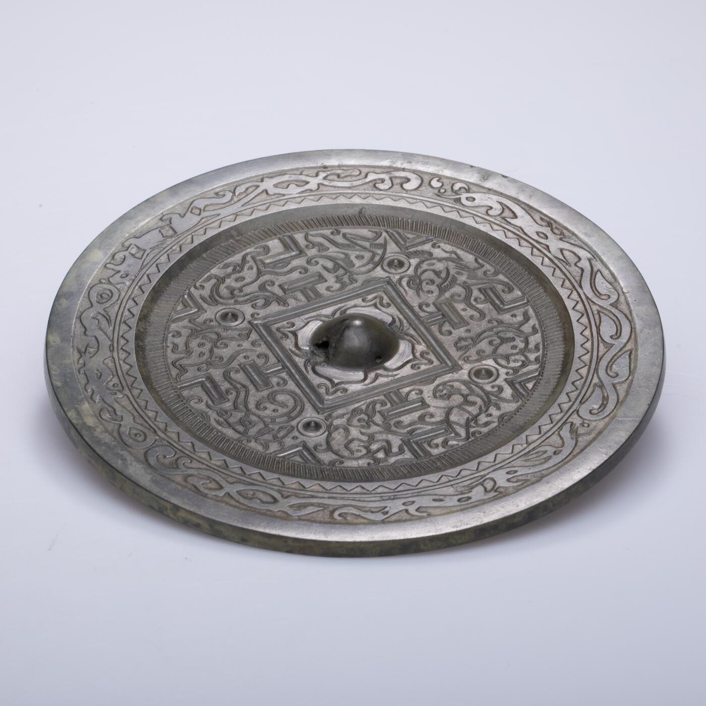 日本大阪雅和auction 2025金秋競賣會第一期 漢 走獸紋規矩鏡 尺寸:D:14cm 備註:盒付   此面漢代走獸紋規矩鏡，鏡體渾圓厚重，通體黑漆古包漿，色澤沉潤柔和，鏡背以精細線條鑄出走獸紋飾，或奔或躍，神態生動，間以幾何規矩紋相間佈局，構圖嚴謹而富節奏感。鏡紐高起，形制端正，邊廓勻整，展現漢代青銅鏡工藝的成熟與規範之美。此類鏡多寓意驅邪護身、吉祥長安，包漿溫潤，光澤深邃，鏡面保存完好，實屬難得。其兼具實用與審美價值，彰顯漢代金屬藝術的莊重典