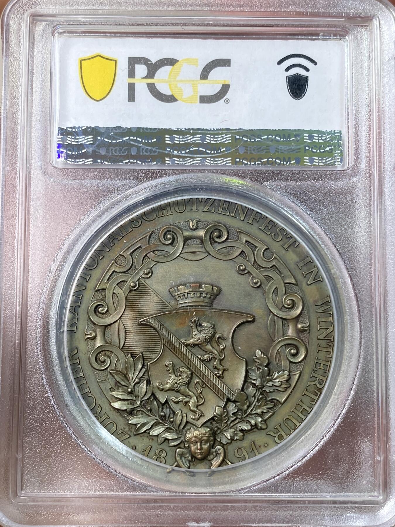 万国钱币拍卖第056期 PCGS SP64 1891年瑞士苏黎世铜章 温特图尔射击节 高浮雕加厚盒 正面:指挥官手握瑞士国旗，指向前方，放箭放箭～跪着的战士正用十字弓瞄准，背景是山地风景。背面:温特图尔的加冕城市纹章，长有翅膀的小天使头像，周围装饰有花环。高分顶级状态，设计极佳，实物非常漂亮。大尺寸铜章，新评几乎都是超大盒子，此枚收纳观赏更为合适。
