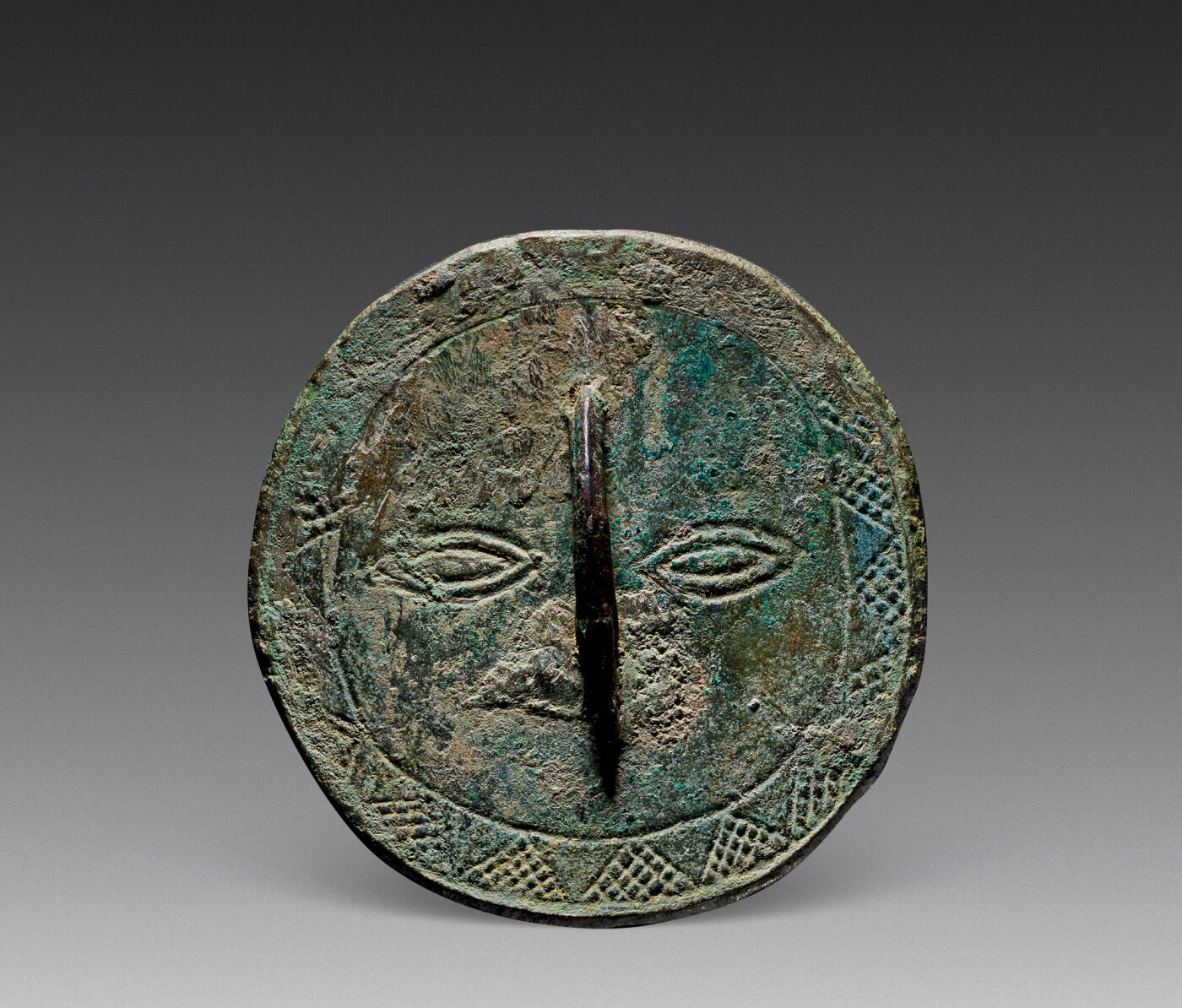 上海匡时2025年秋季拍卖会 中国历代钱币 铜镜专场 太阳神纹镜 不规则圆形，鼻形钮。主纹饰为造型古朴的一张人面纹，圆脸凤眼，双目炯炯有神，桥钮为鼻，钮下纹饰为嘴，外围两周圈带中间饰锯齿纹，形成太阳光芒纹，其外填满鱼子点纹。若作太阳崇拜理解时，人脸即是太阳神，锯齿应系太阳光芒，鱼子点似为「满天星星」。整个人脸呆萌天真，造型抽象又形象，具有鲜明的地域与时代特征，品种极为少见，此枚纹饰仅见，包浆熟润，保存完整。来源：台北“一雅堂”旧藏。参