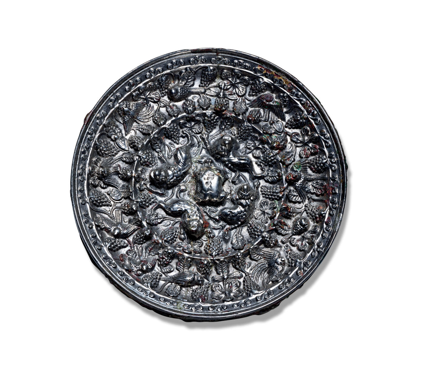 上海匡时2025年秋季拍卖会 中国历代钱币 铜镜专场 海兽葡萄镜 圆形，伏兽钮。镜背空间由一周凸棱划分为内外两区。内区环绕镜钮饰有四只昂首趴伏的海兽，形体饱满，刻画精细，姿态憨拙可掬；其间满铺细密交错的葡萄果实与枝蔓，枝蔓延伸跨越凸梁进入外区，形成所谓“过梁葡萄”的独特构图。外区在繁茂的葡萄藤蔓间点缀四只展翅飞翔的禽鸟，动静相宜，生意盎然，俨然一幅充满生趣的乐园图景。镜缘饰有一周卷叶纹。