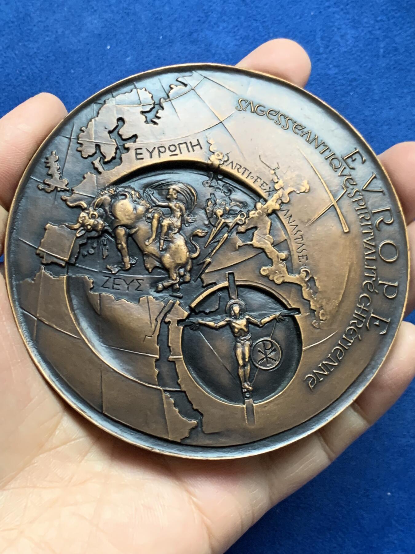 《竞宝斋》第452场 周日，周一 2场连拍 （全场包邮，欢迎送拍） 名家名作 德梅特尔·阿纳斯塔斯设计 迈向欧洲人文主义 超大号纪念铜章 506.5克，96.5mm，丰饶角铜标，此为原铸章， 市场出现大部分为边铭处带年份的后铸章  正面： 在地球仪上，描绘了宙斯化身为公牛掳走欧罗巴仙女的传说场景；下方圆形图案中为十字架上的基督及其花押字   背面：在欧洲地图上，三位分别代表地下、陆地与太空领域的主宰者。圆形图案中，两人在象征欧洲的树下握手，下方是希望之舟。