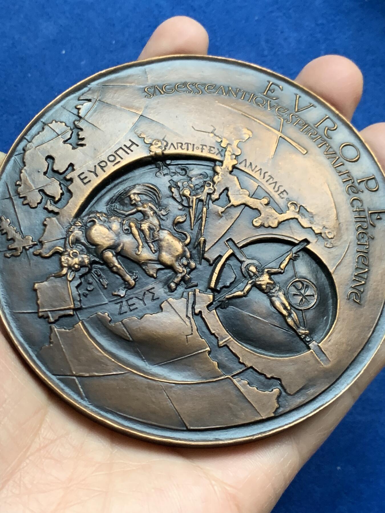 《竞宝斋》第452场 周日，周一 2场连拍 （全场包邮，欢迎送拍） 名家名作 德梅特尔·阿纳斯塔斯设计 迈向欧洲人文主义 超大号纪念铜章 506.5克，96.5mm，丰饶角铜标，此为原铸章， 市场出现大部分为边铭处带年份的后铸章  正面： 在地球仪上，描绘了宙斯化身为公牛掳走欧罗巴仙女的传说场景；下方圆形图案中为十字架上的基督及其花押字   背面：在欧洲地图上，三位分别代表地下、陆地与太空领域的主宰者。圆形图案中，两人在象征欧洲的树下握手，下方是希望之舟。