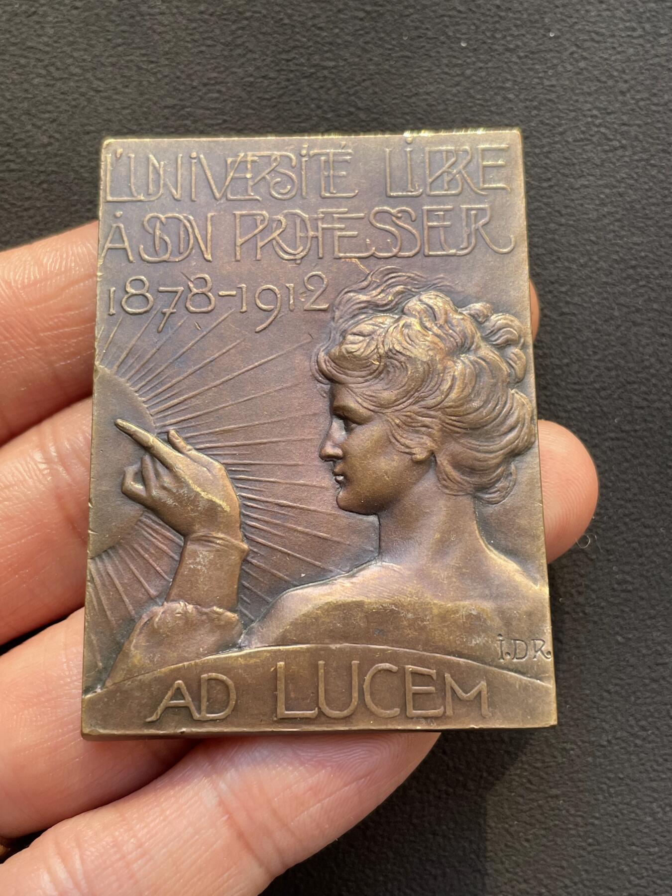 「木头集藏」章牌专场-第65场 比利时布鲁塞尔自由大学教授赫克托·丹尼斯纪念章， 尺寸：59×42mm，81.5g。 赫克托·丹尼斯教授于1878年至1912年间，在自由大学担任教职34年。这枚长方形铜章就是为纪念他的卓越奉献而定制的。 正面是教授的左侧胸像，下方铭文是他的姓名。背面为知识女神点向象征光明的太阳图案，上方三行铭文意为“自由大学”“教授”“1878年至1912年”。