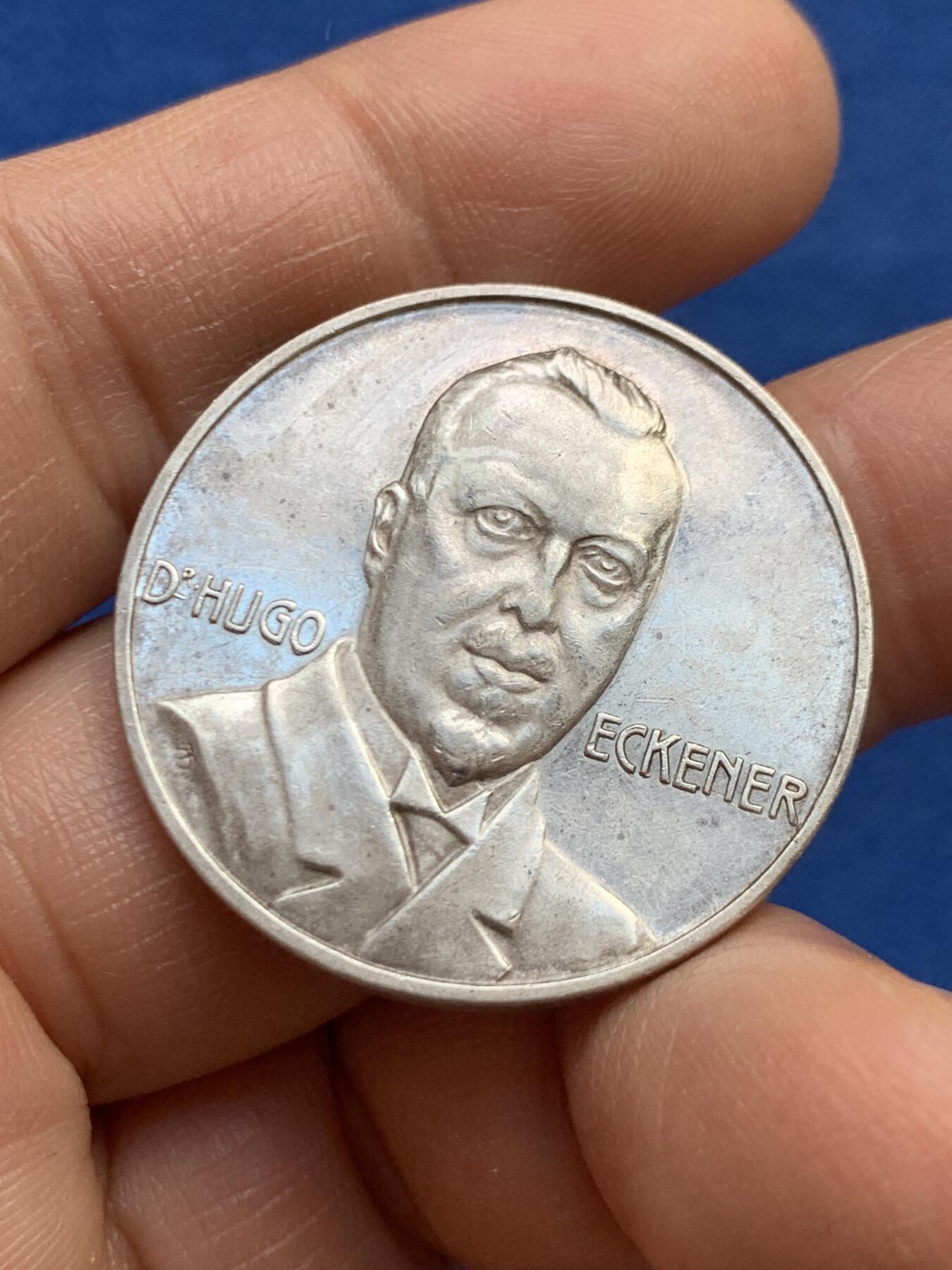 《竞宝斋》第459场 周日，周一，2场连拍 （全场包邮）欢迎送拍 德国雨果·埃克纳纪念银章，他是20世纪初飞艇技术的重要推动者，曾担任齐柏林飞艇公司的负责人，并亲自驾驶“兴登堡号”等著名飞艇完成多次跨大西洋飞行。该章背面详细列出了飞艇的行程时间表，包括从弗里德里希港出发、离开欧洲、经过亚速尔群岛法亚尔、到达萨布尔岛、波士顿、纽约和莱克赫斯特的具体日期和时间。14.92克，33.3mm，带990银标。