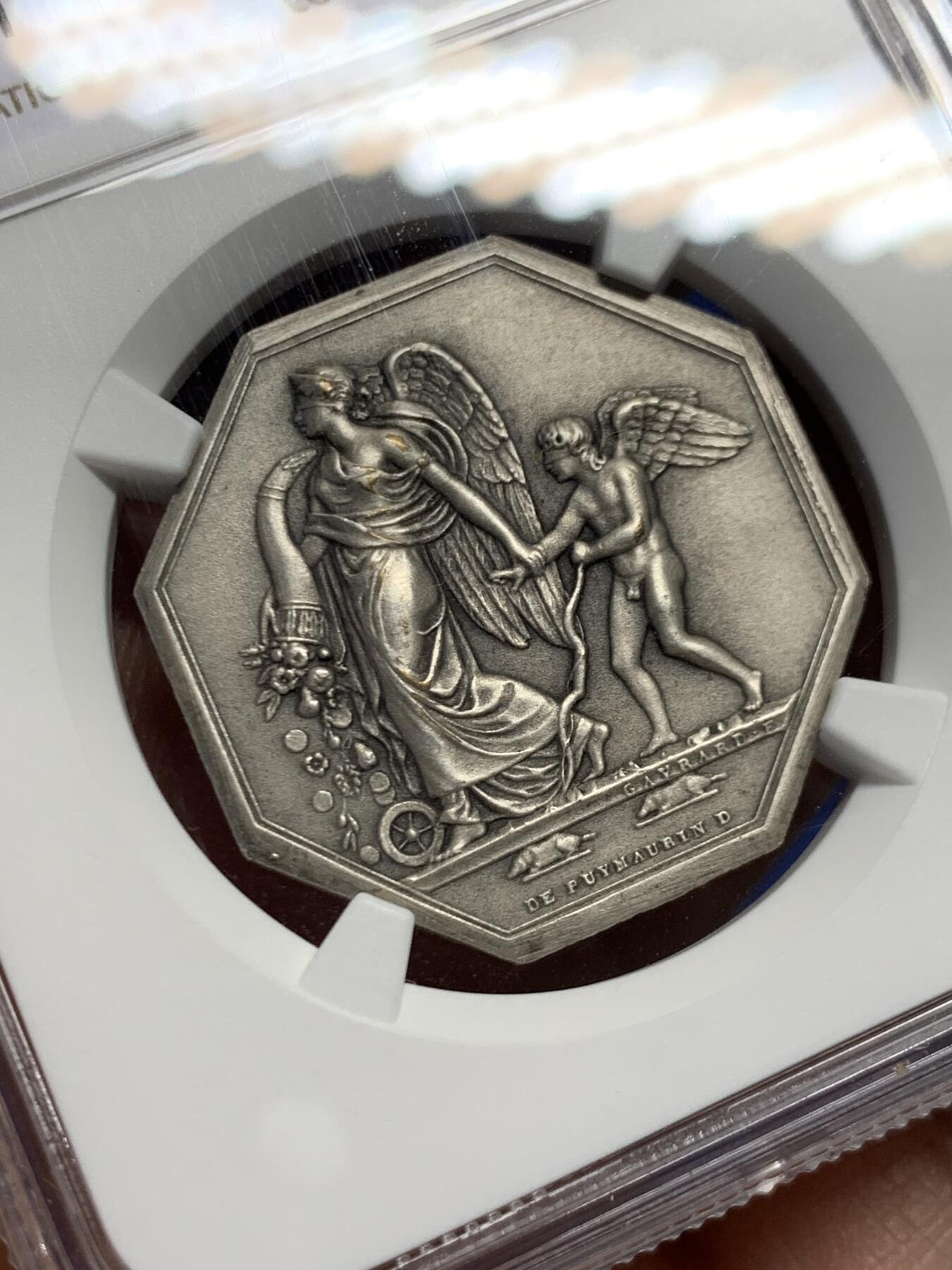 《竞宝斋》第460场 周日，周一，2场连拍 （全场包邮）欢迎送拍 NGC MS63 法国八角镀银铜质代币，正面雕刻一位羽翼女神(胜利或命运女神)牵引着小天使(爱神丘比特)，象征着命运引导下的爱与希望，左侧为撒落的丰收之角(Cornucopia)，右下角有艺术家名“DE PUYMAURIN”与雕刻标记，与整体构图共同形成寓言式场景。