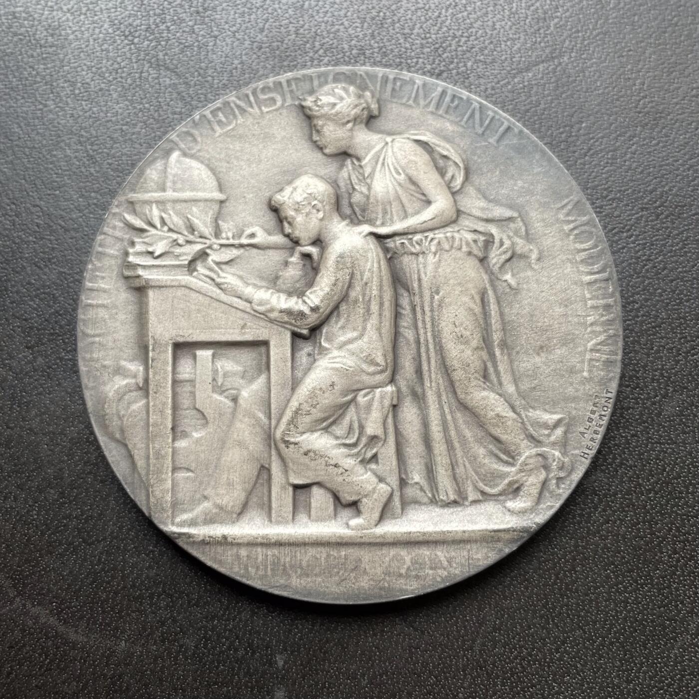 「木头集藏」章牌精品专场！ Albert Herbemont～法国现代教育协会银章 ，直径：50 mm，64 g，边铭：丰饶角 1ARGENT。少见的银章品种。正面女神把橄榄枝放在学生的书桌上，背面旭日东升，橡树枝繁叶茂。表面采用磨砂工艺，有一些氧化形成的深色纹路，不是划痕，实物品相很好。