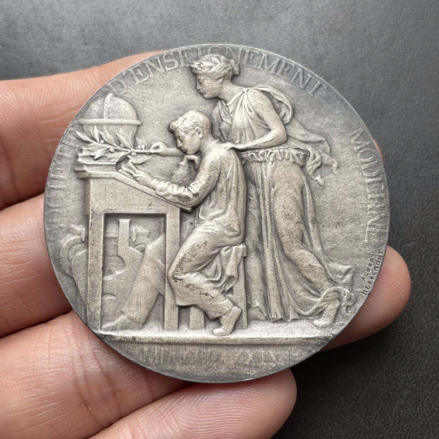 「木头集藏」章牌精品专场！ Albert Herbemont～法国现代教育协会银章 ，直径：50 mm，64 g，边铭：丰饶角 1ARGENT。少见的银章品种。正面女神把橄榄枝放在学生的书桌上，背面旭日东升，橡树枝繁叶茂。表面采用磨砂工艺，有一些氧化形成的深色纹路，不是划痕，实物品相很好。
