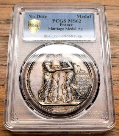 凡希社世界钱币微拍第三百十九期 - 十九世纪末法国天主教婚姻纪念银章PCGS-MS62，正面是一对古典风格的男女牵手而立，身旁有小狗相伴，背景为砖墙，象征婚姻的陪伴与安稳生活，整体浮雕细腻，充满浪漫主义风格。 背面中央是花押式的字母组合，外围环绕玫瑰花环，象征爱情与婚姻的美好，是当时定制婚礼纪念章的典型布局。