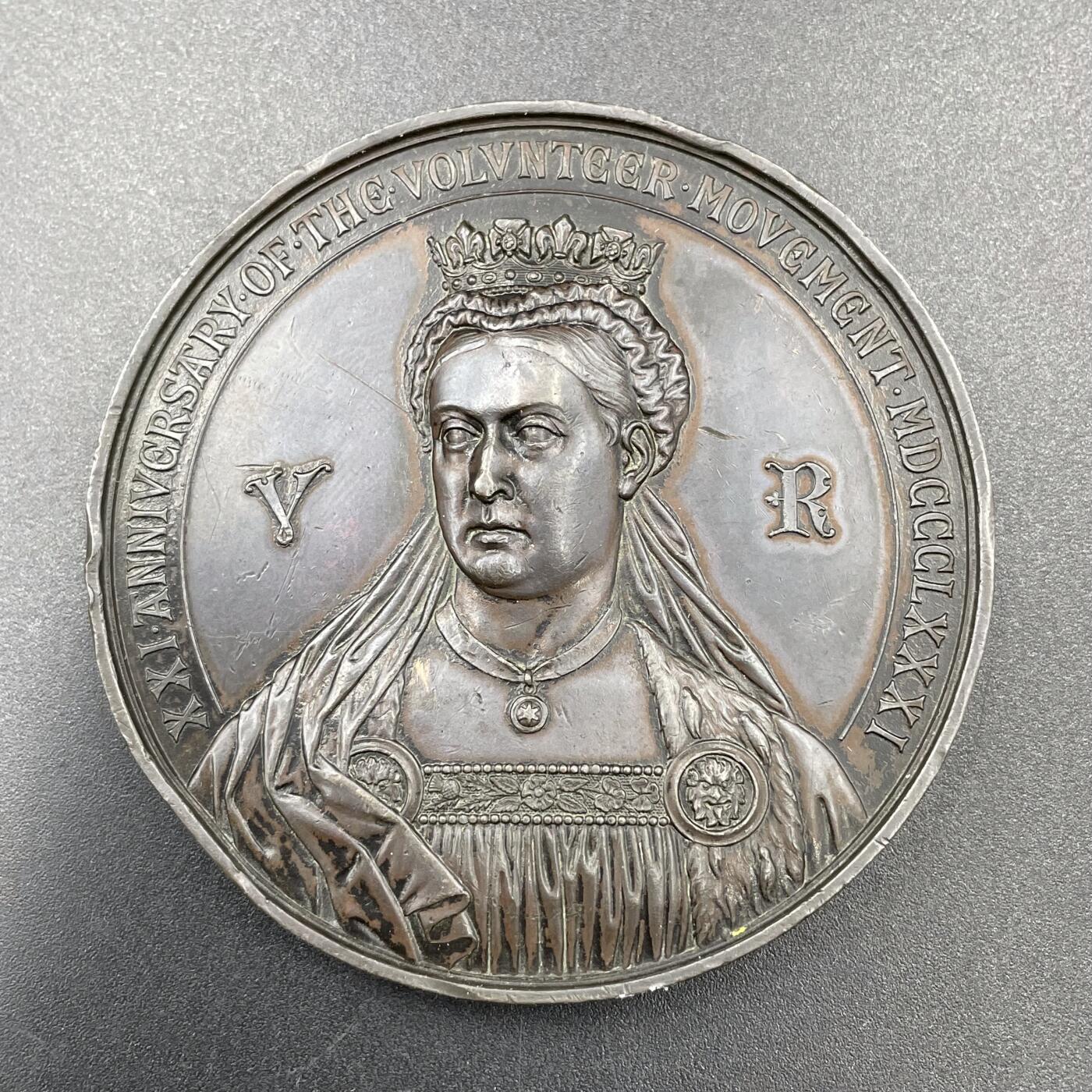 「木头集藏」章牌专场-第67场 1881年英国志愿兵运动21周年纪念铜章，直径：63.5mm，93.8 g，中央刻画天使长米迦勒（St. Michael），他身披盔甲、手持盾牌与宝剑，象征着守护与勇气。左侧是怀抱孩子的母亲与少女，手持橄榄枝，代表家园与和平；右侧是跪姿的志愿兵与列队士兵，背景还有帆船，呼应了志愿兵海陆防御的职责。