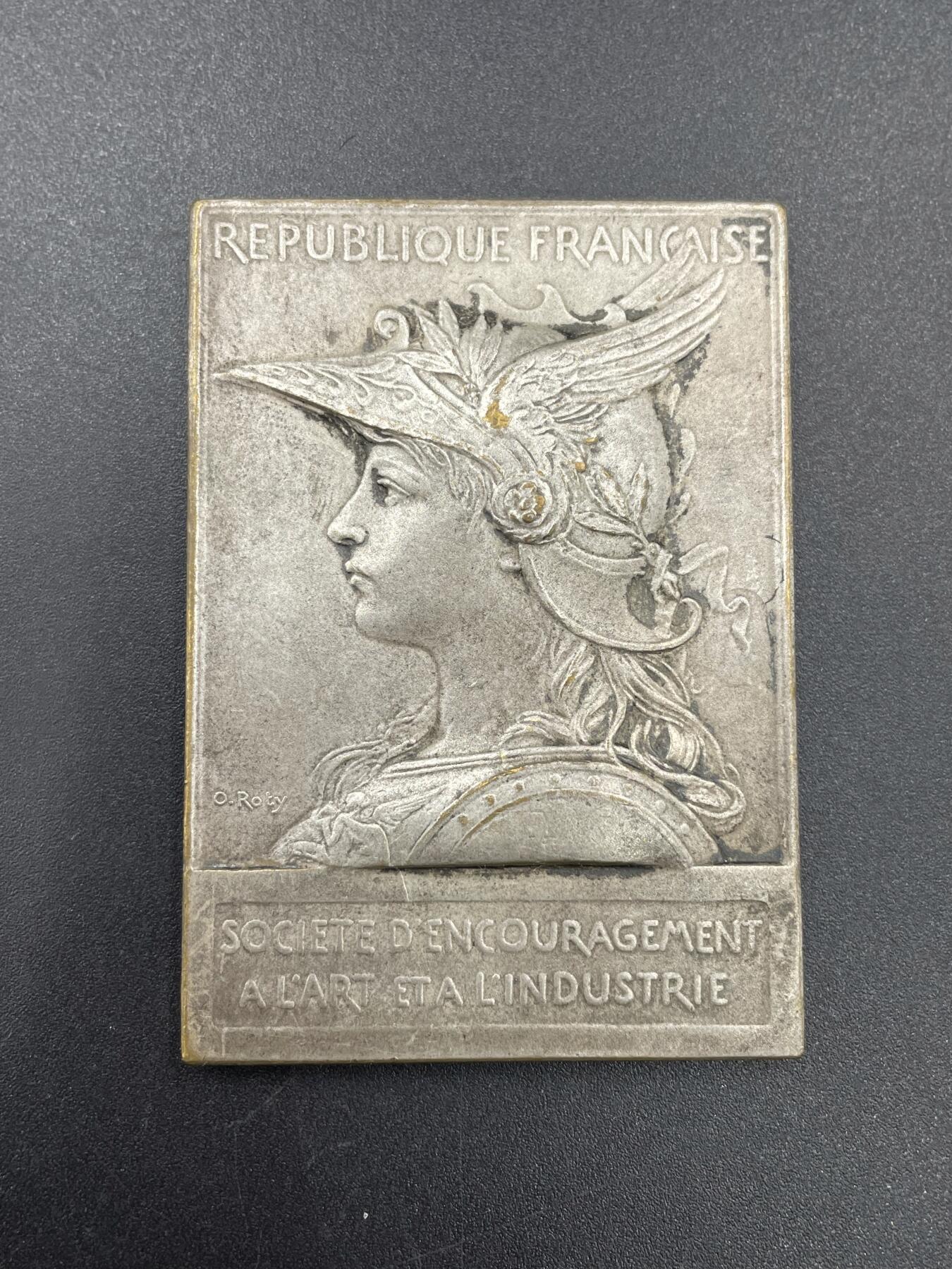 「木头集藏」章牌专场-第67场 O.Roty～法国艺术与工业促进会镀银章，尺寸：42 × 60 mm ，73 g，边铭：丰饶角 BRONZE， 此章正面采用了罗蒂经典的戎装玛丽安娜头像。上方是法语“法兰西共和国”，下方是“艺术与工业促进会”铭文。背面图案为古希腊神话中掌管火、锻造与工匠技艺的赫菲斯托斯，在向智慧女神雅典娜展示将美杜莎头颅镶嵌上去的盾牌。上方铭文