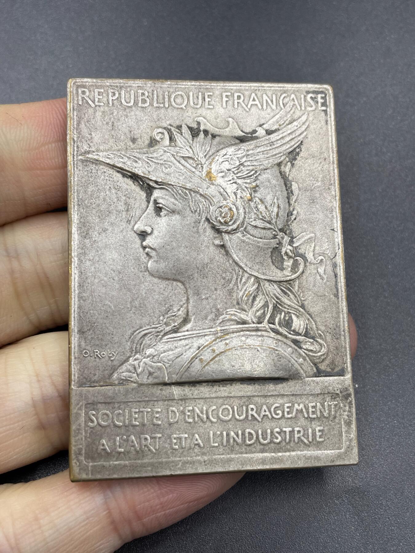 「木头集藏」章牌专场-第67场 O.Roty～法国艺术与工业促进会镀银章，尺寸：42 × 60 mm ，73 g，边铭：丰饶角 BRONZE， 此章正面采用了罗蒂经典的戎装玛丽安娜头像。上方是法语“法兰西共和国”，下方是“艺术与工业促进会”铭文。背面图案为古希腊神话中掌管火、锻造与工匠技艺的赫菲斯托斯，在向智慧女神雅典娜展示将美杜莎头颅镶嵌上去的盾牌。上方铭文