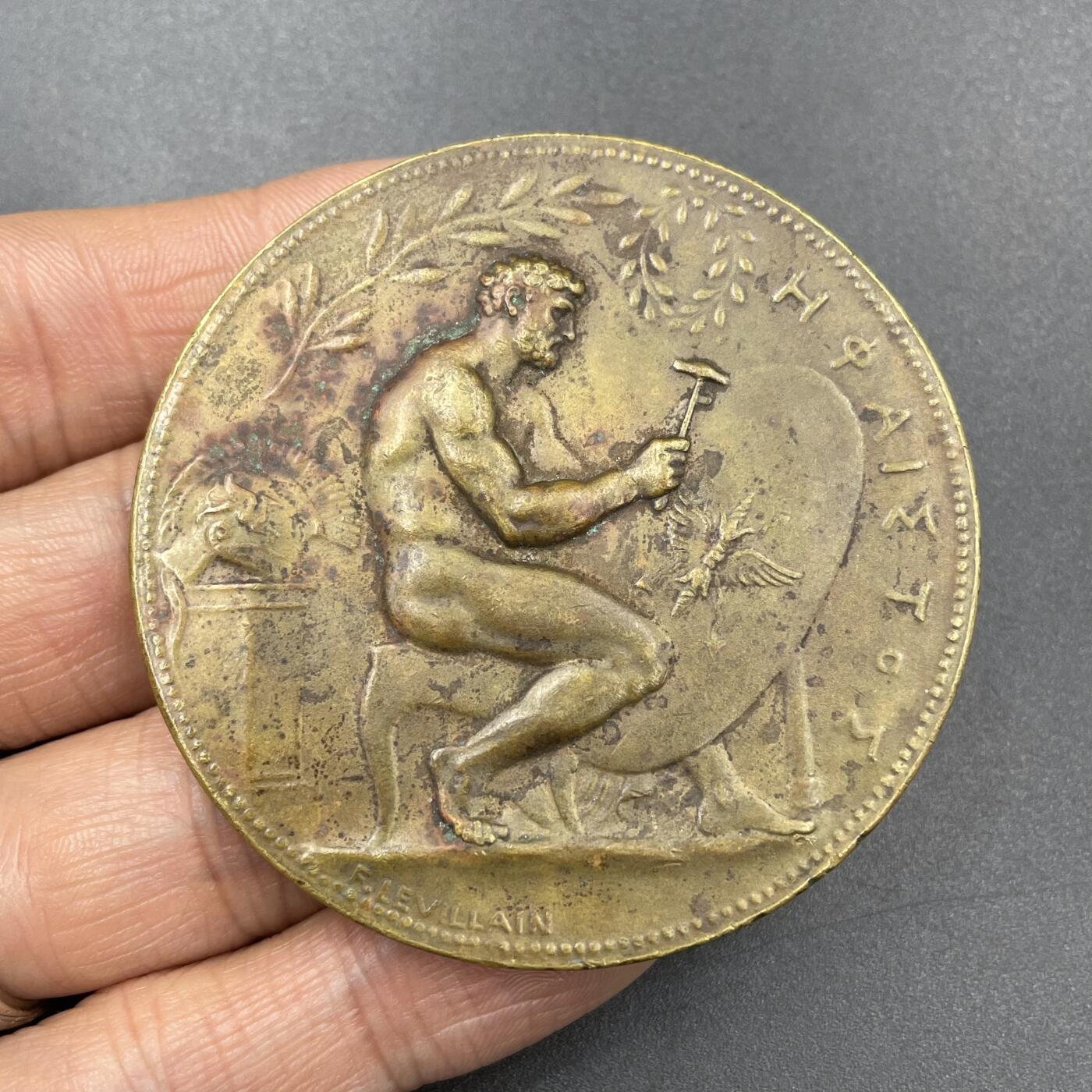 「木头集藏」章牌专场-第67场 F Levillain～1888年法国火神赫淮斯托斯为宙斯打造盾牌铜章 ，直径：58mm，边铭：丰饶角 BRONZE，马耶尔目录（1870-1940年法国章牌艺术）126号作品。背面铭文显示此章是巴黎青铜制造商协会颁发给绘画比赛并列第一名的P. Lebrun。正面是正在打造宙斯盾牌的赫淮斯托斯。赫淮斯托斯是宙斯和赫拉的儿子，希腊神话中的火神和匠神，为诸神打造了很多兵器。