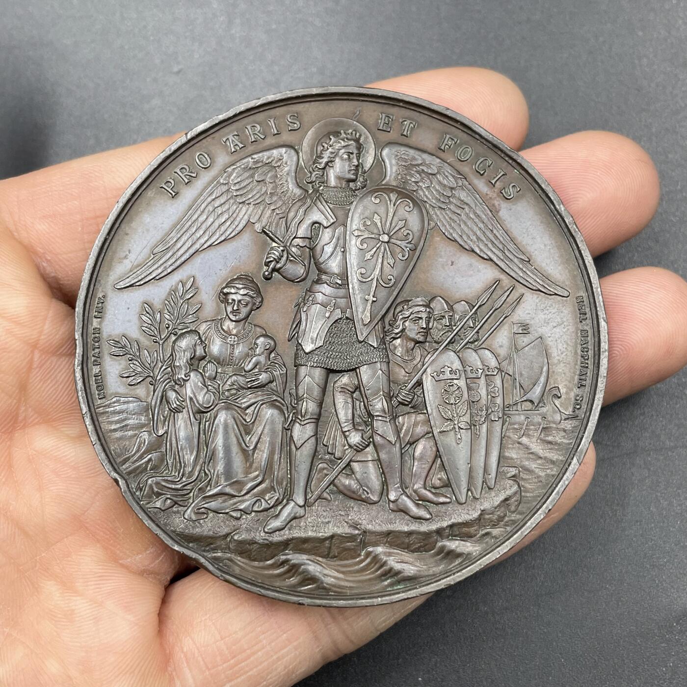 「木头集藏」章牌专场-第67场 1881年英国志愿兵运动21周年纪念铜章，直径：63.5mm，93.8 g，中央刻画天使长米迦勒（St. Michael），他身披盔甲、手持盾牌与宝剑，象征着守护与勇气。左侧是怀抱孩子的母亲与少女，手持橄榄枝，代表家园与和平；右侧是跪姿的志愿兵与列队士兵，背景还有帆船，呼应了志愿兵海陆防御的职责。