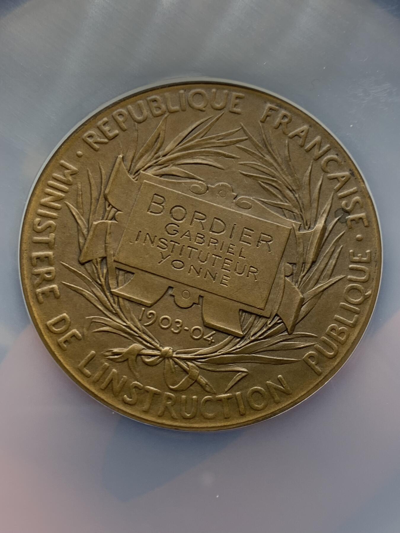 《竞宝斋》第465场 周日，周一，周二，3场连拍 （全场包邮）欢迎送拍 NGC MS64BN 1904年法国初级教育铜章，罗蒂作品，直径51毫米，正面图案展现了一位女性教师向两名学生传授知识的场景，象征着国家对基础教育的重视。加大盒