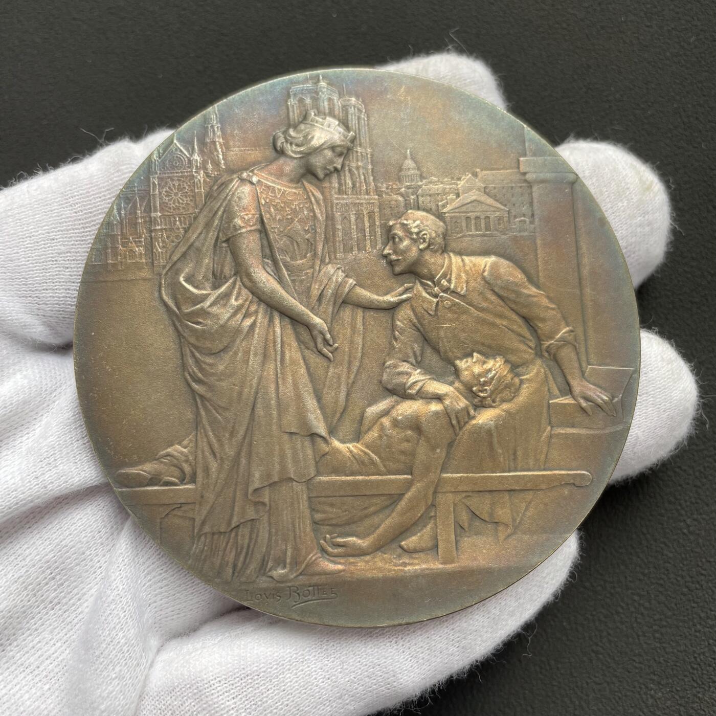 「木头集藏」“ 新岁撷珍”——章牌精品专场！  Louis Bottée～法国巴黎市民医院内科与外科住院医师制度百年纪念镀银大铜章，直径：68 mm，124.5 g，这枚章属于欧洲新艺术风格章牌的经典品种，兼具艺术价值与历史纪念意义。正面：描绘了象征医学的女性形象，俯身关怀病床旁的医生与患者，寓意医学的人文关怀。背景是巴黎圣母院。