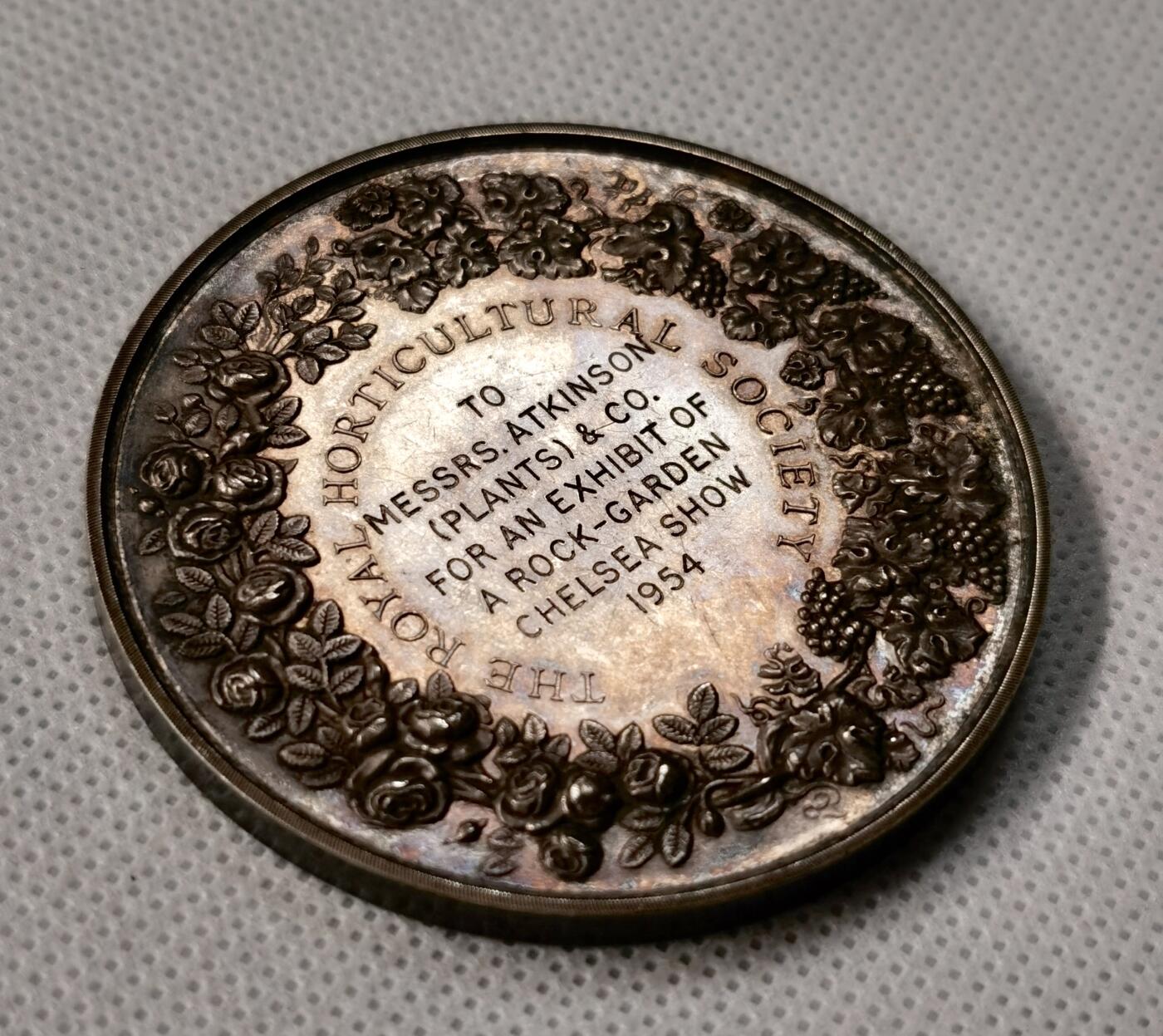 “福币呈祥”新年甄品场（直播拍卖）甄臻铺-世界币章拍卖（第50期），送拍享返佣金等优惠多多 1954年英国皇家园艺学会（Royal Horticultural Society）银质奖章，直径55毫米，重70克，浮雕工艺精细，正面为古典女神像，背面环绕花卉和英文铭文，具体见图。奖章为1954年切尔西花展（Chelsea Show）颁发，授予Messrs. Atkinson（Plants）& Co.的岩园展示，历史背景清晰，品相包浆几近完美