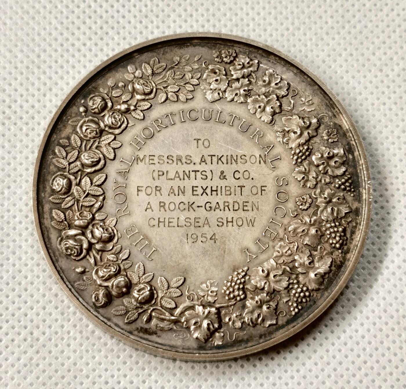 “福币呈祥”新年甄品场（直播拍卖）甄臻铺-世界币章拍卖（第50期），送拍享返佣金等优惠多多 1954年英国皇家园艺学会（Royal Horticultural Society）银质奖章，直径55毫米，重70克，浮雕工艺精细，正面为古典女神像，背面环绕花卉和英文铭文，具体见图。奖章为1954年切尔西花展（Chelsea Show）颁发，授予Messrs. Atkinson（Plants）& Co.的岩园展示，历史背景清晰，品相包浆几近完美