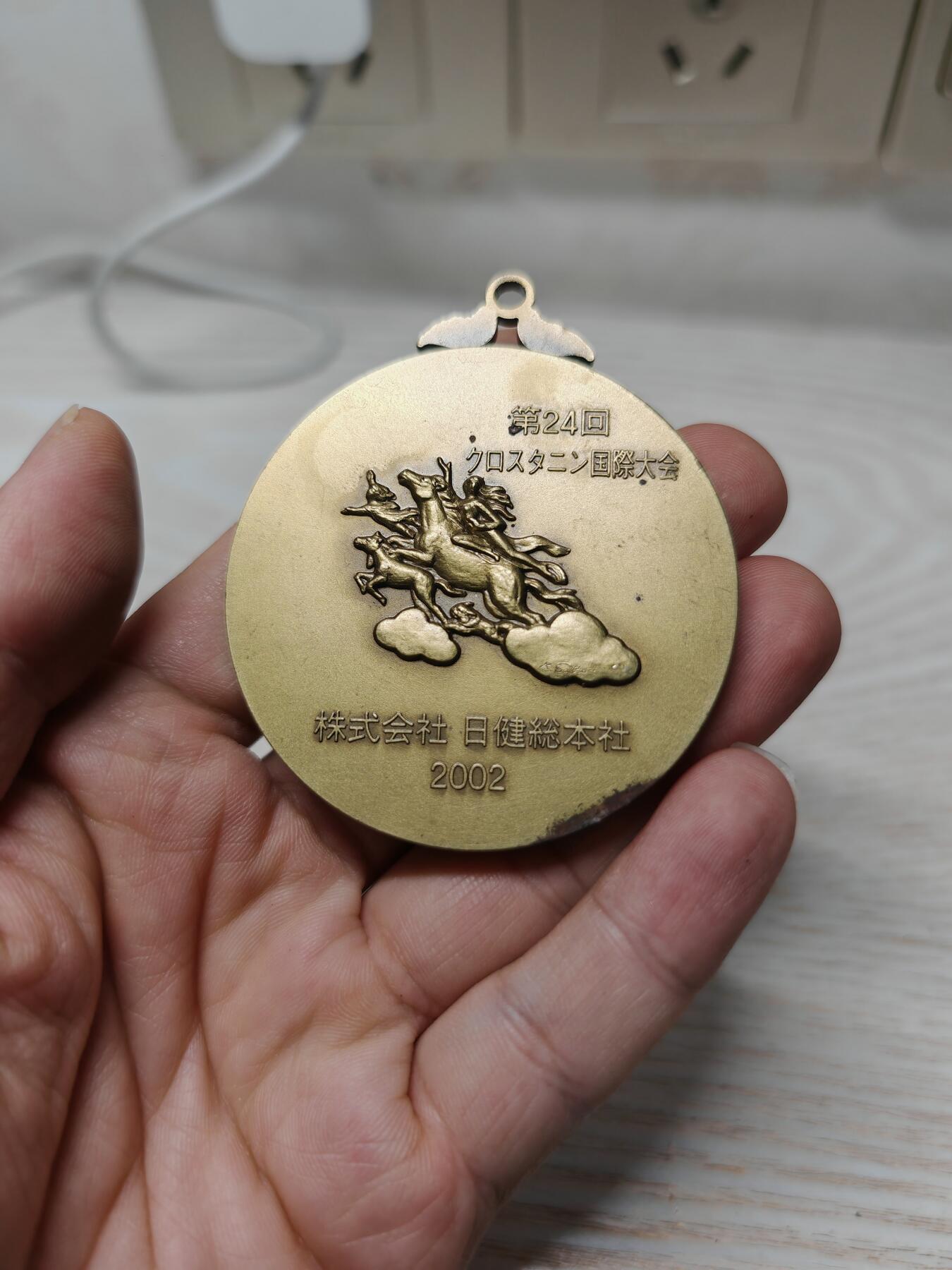 2002年第24届克罗斯塔宁国际大会纪念奖牌人物立体 - 正面：浮雕为日健総本社的创始人田中美穂的头像，右下角有雕刻师签名。 - 背面：刻有“第24回クロスタニン国際大会”、“株式会社 日健総本社”和年份“2002”，并配有飞马与人物的神话风格浮雕。 浮雕工艺精细，背面的飞马神话图案设计独特，具有一定的艺术观赏性。