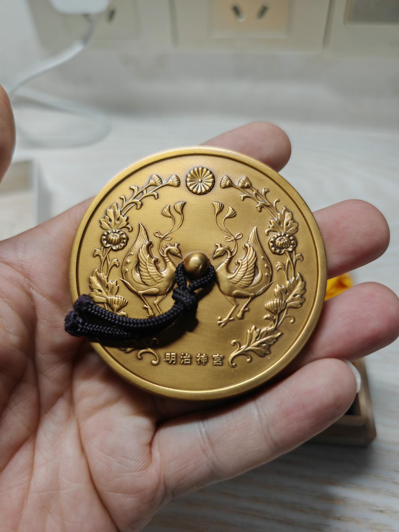 日本明治神宮的铜制纪念章 中央是双凤凰相对而立，周围环绕着菊花、牡丹等花卉纹样，顶部是日本皇室的十六瓣八重表菊纹。 背面是明治天皇的御制和歌，大意是：“在您的光辉下，朝阳格外耀眼；为了您，我心怀清爽而思念。