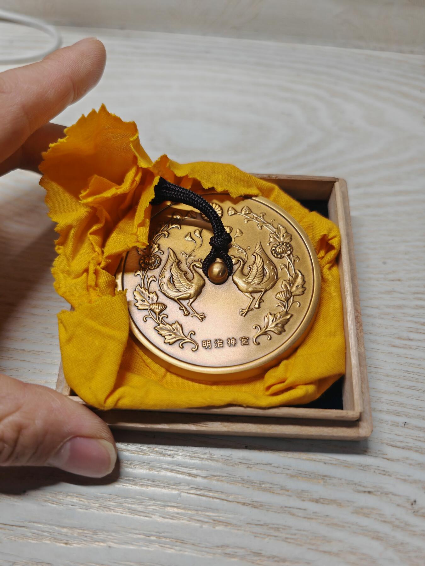 日本明治神宮的铜制纪念章 中央是双凤凰相对而立，周围环绕着菊花、牡丹等花卉纹样，顶部是日本皇室的十六瓣八重表菊纹。 背面是明治天皇的御制和歌，大意是：“在您的光辉下，朝阳格外耀眼；为了您，我心怀清爽而思念。