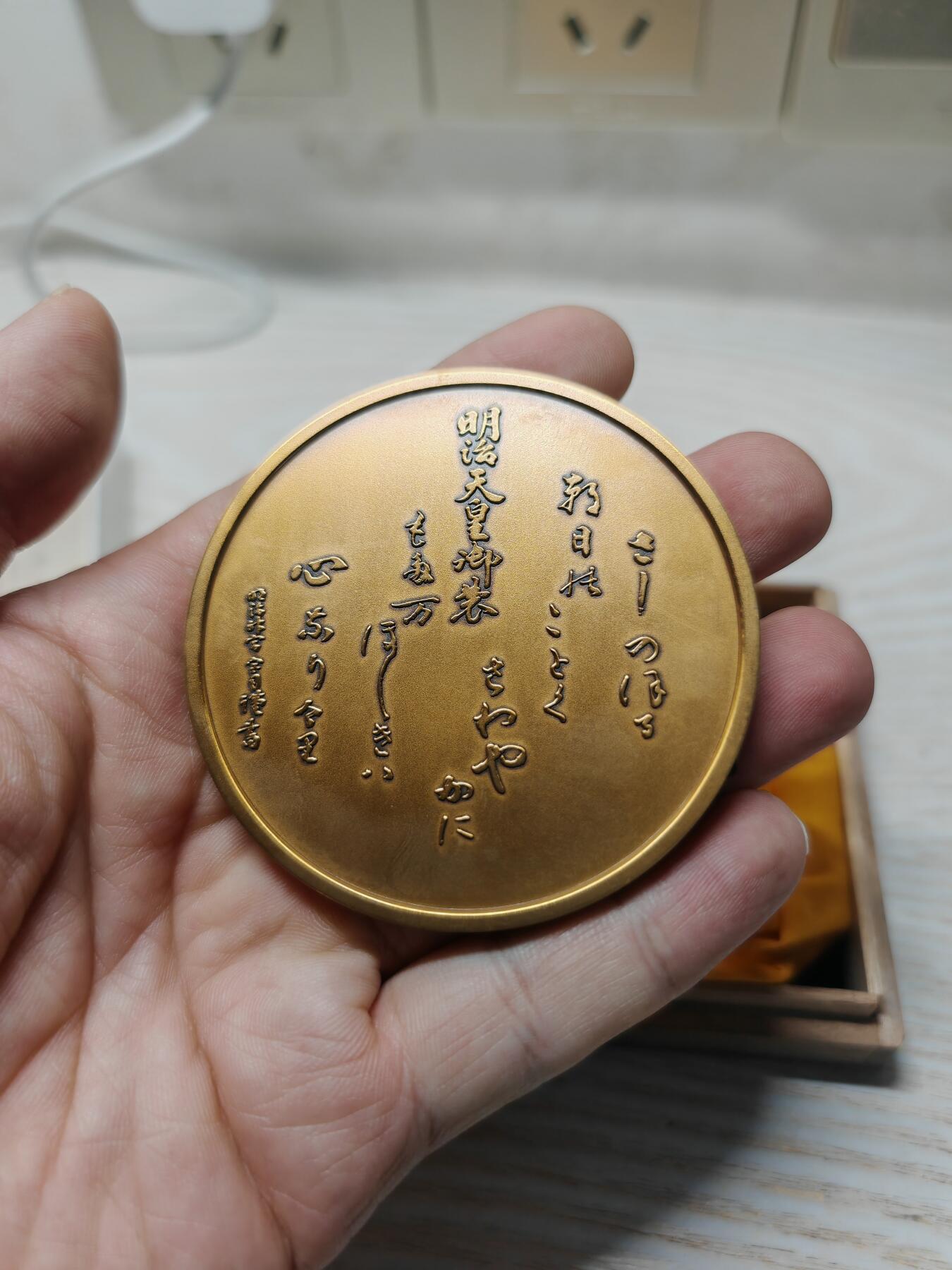 日本明治神宮的铜制纪念章 中央是双凤凰相对而立，周围环绕着菊花、牡丹等花卉纹样，顶部是日本皇室的十六瓣八重表菊纹。 背面是明治天皇的御制和歌，大意是：“在您的光辉下，朝阳格外耀眼；为了您，我心怀清爽而思念。