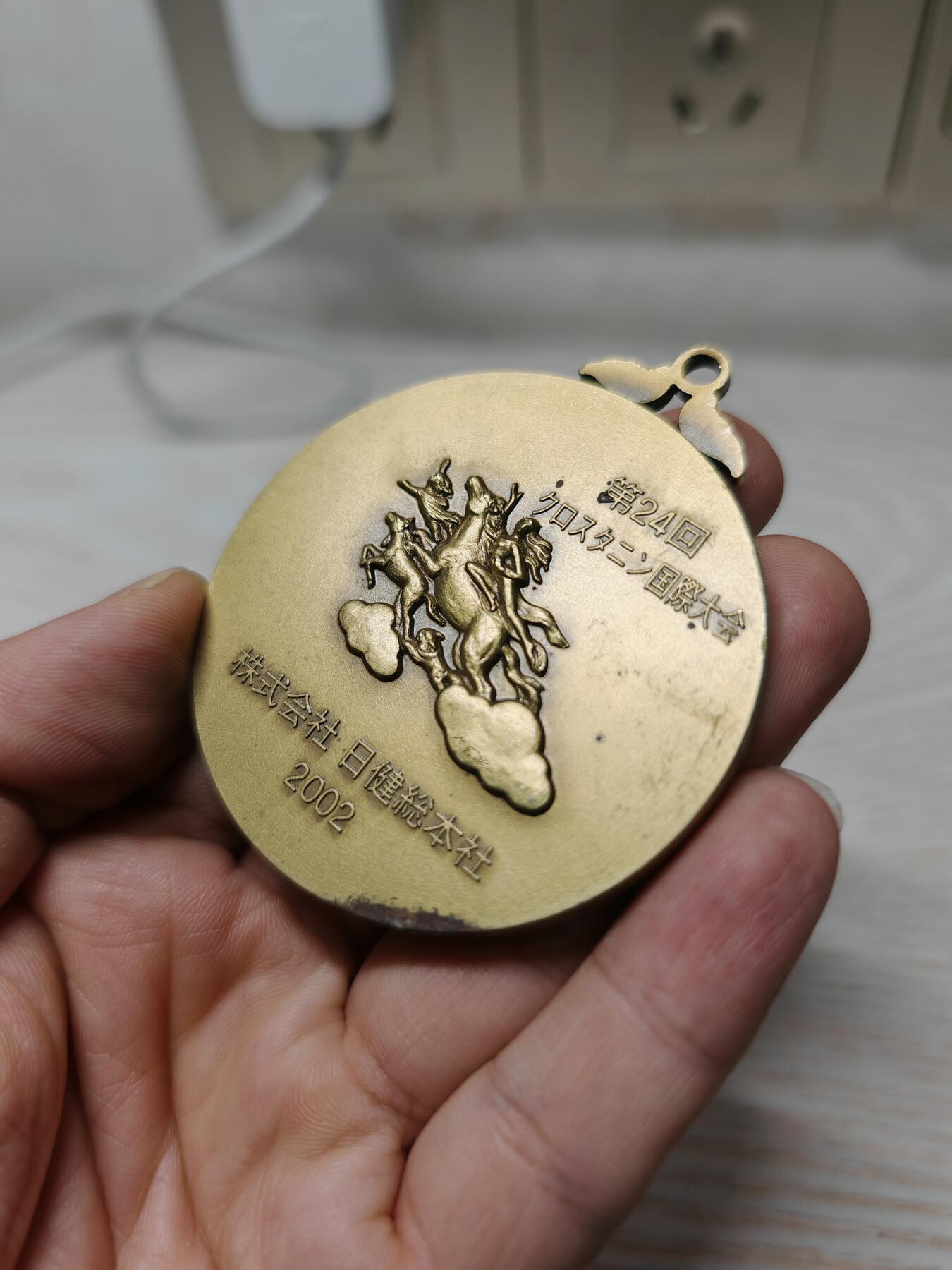 2002年第24届克罗斯塔宁国际大会纪念奖牌人物立体 - 正面：浮雕为日健総本社的创始人田中美穂的头像，右下角有雕刻师签名。 - 背面：刻有“第24回クロスタニン国際大会”、“株式会社 日健総本社”和年份“2002”，并配有飞马与人物的神话风格浮雕。 浮雕工艺精细，背面的飞马神话图案设计独特，具有一定的艺术观赏性。