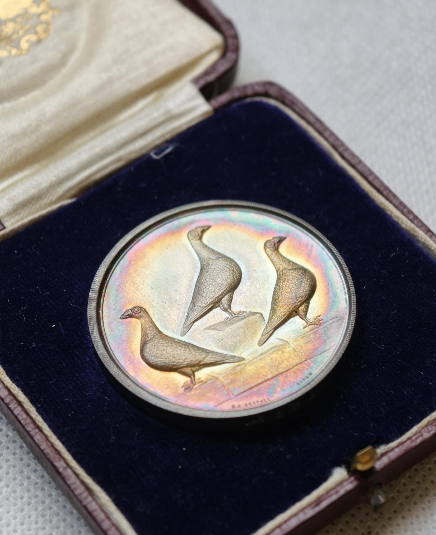 甄臻铺-世界币章拍卖（第54期），3月28日甄品场拍品征集中，送拍享返佣金等优惠多多 1927年英国龙骑士赛鸽俱乐部获奖银章，直径39毫米，重32克。正面雕刻三只鸽子图案，背面刻有W.H.Inman获奖字样。原装红皮盒，带内衬丝绒垫。表面自然氧化呈现五彩包浆，工艺精湛。边缘清晰，铭文完整，原装盒子完整（鸽子重拍）