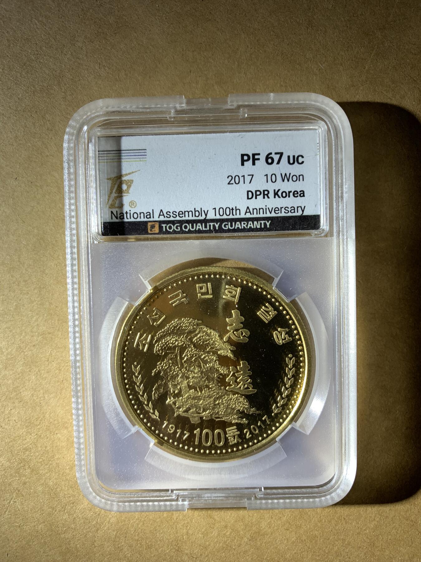【礼羽收藏】🌏世界钱币拍卖第132期 朝鲜2017年发行的10元纪念币，纪念最高人民会议成立100周年，评级PF 67 UC（精制币，镜面深打67分）。材质是铜镍合金，金黄色调，正面有“志远”和松树图案。