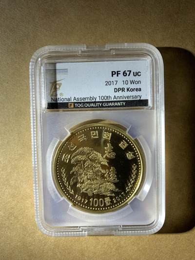 【礼羽收藏】🌏世界钱币拍卖第132期 - 朝鲜2017年发行的10元纪念币，纪念最高人民会议成立100周年，评级PF 67 UC（精制币，镜面深打67分）。材质是铜镍合金，金黄色调，正面有“志远”和松树图案。