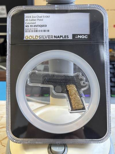 第219期拍卖 - 乍得2024年美国M1911手枪2盎司异形纪念银币NGC-MS70，999银，2盎司，发行1000枚，评级盒有破.整币复刻 M1911 手枪轮廓，枪身黑色仿古做旧，握把位置镀金浮雕自由女神图案，异形特殊封装盒。异形手枪形制，**Colorized（彩印）+Antiqued 仿古做旧 + 局部镀金